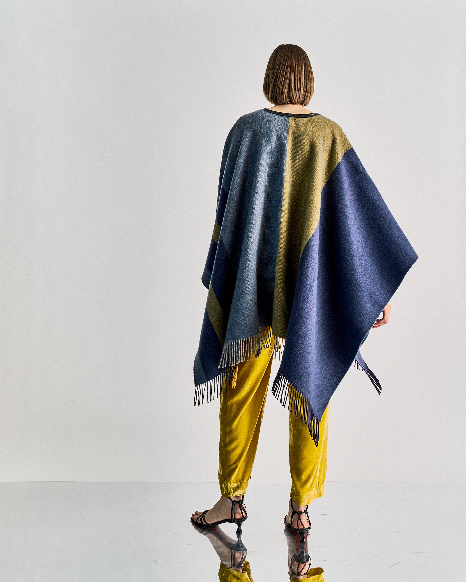 A person stands facing away, wearing a blue and fringed Hermès poncho over bright yellow pants and black sandals. 