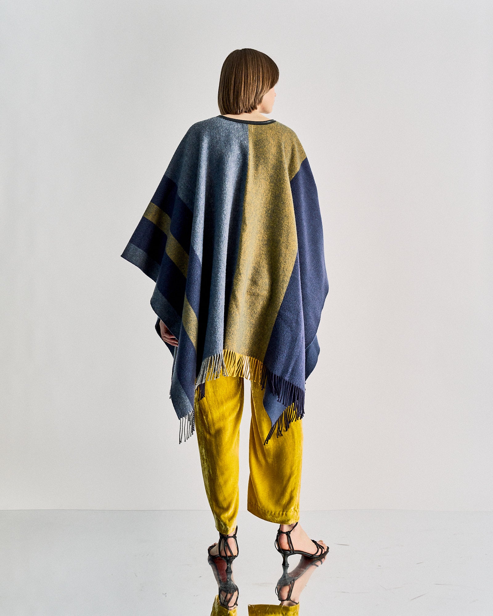 A person stands facing away, wearing a blue and fringed Hermès poncho over bright yellow velvet pants and black sandals. 