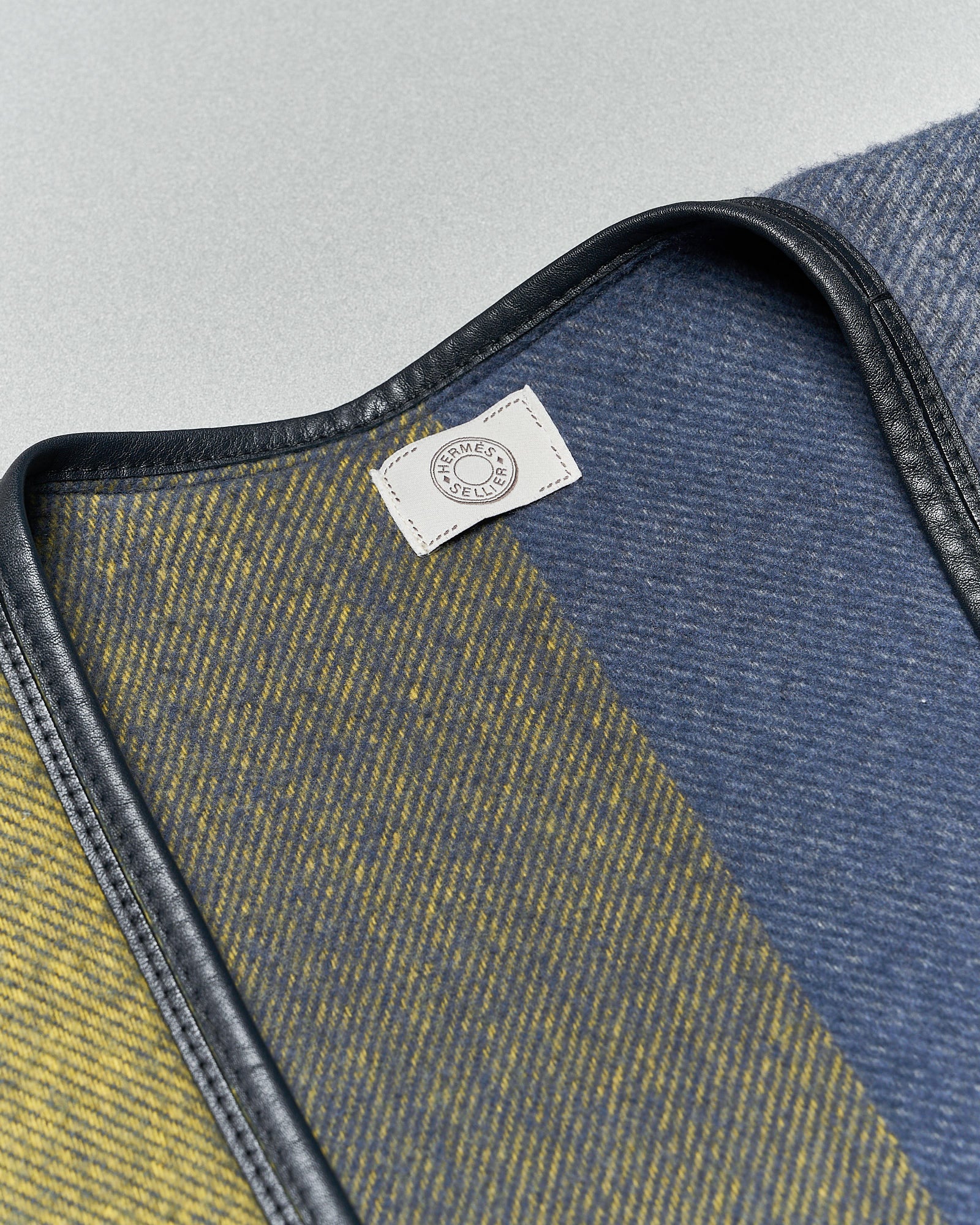 Close-up of a fabric with diagonal stripes in mustard and navy shades, bordered by black leather trim. A small white label is attached.