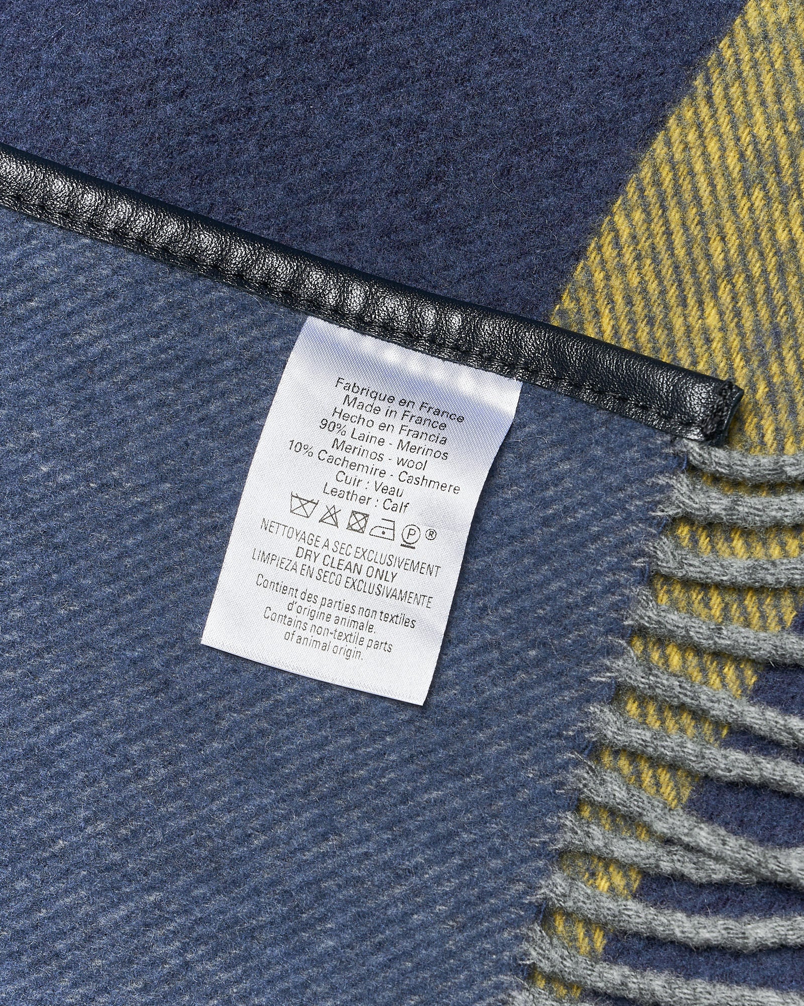 Close-up of a fabric label on a blue and yellow wool blanket. The label indicates it's made in France with merino, cashmere, and calf leather.