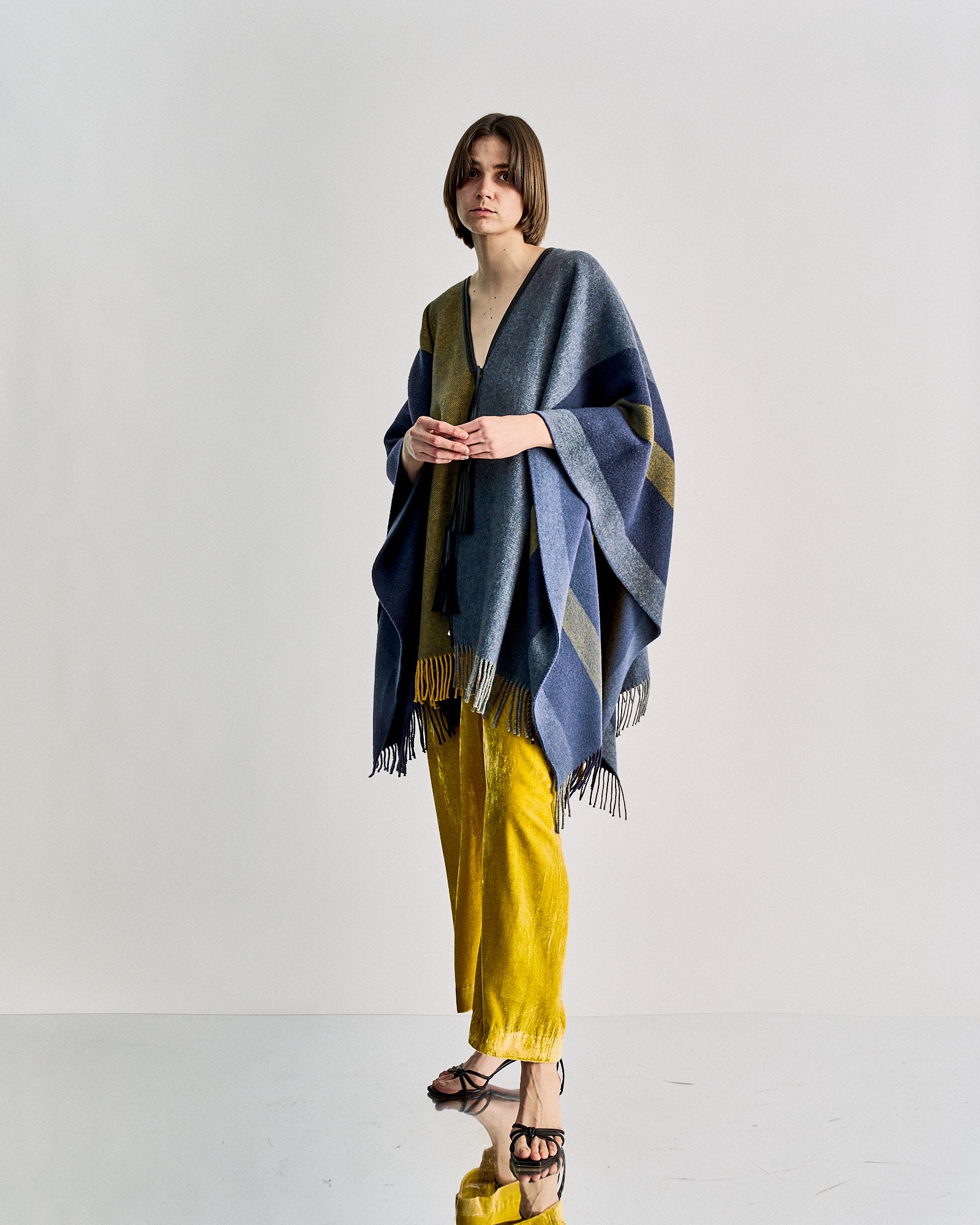 A person wears a blue Hermès poncho with stripes and mustard yellow velvet pants, standing against a light background. 