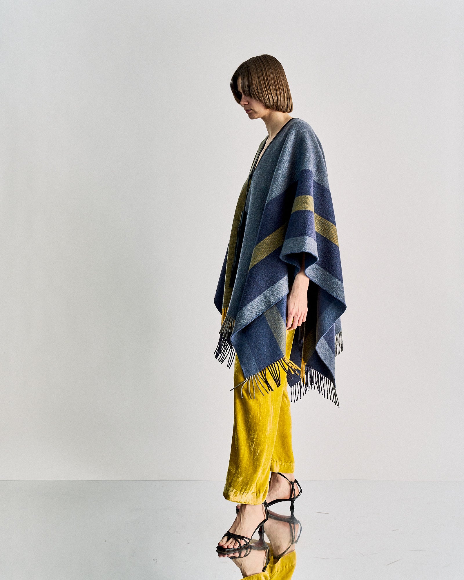 A person in a colourful Hermès poncho with blue and mustard stripes stands sideways against a plain background. They wear yellow pants and strappy black sandals.