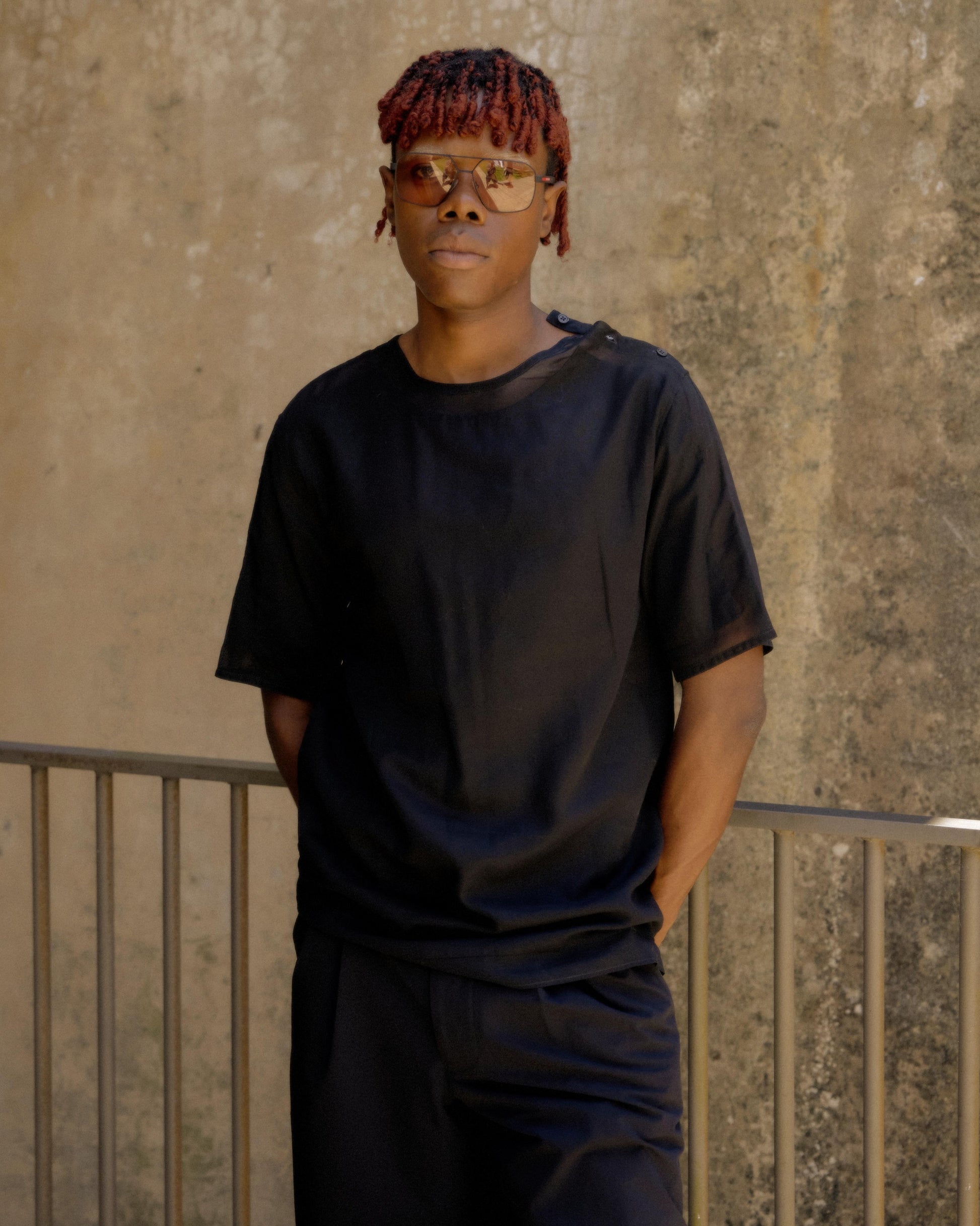 A person wearing a black sheer Hermes t-shirt with three button closures at the neck, standing in a casual pose with one hand in a pocket.