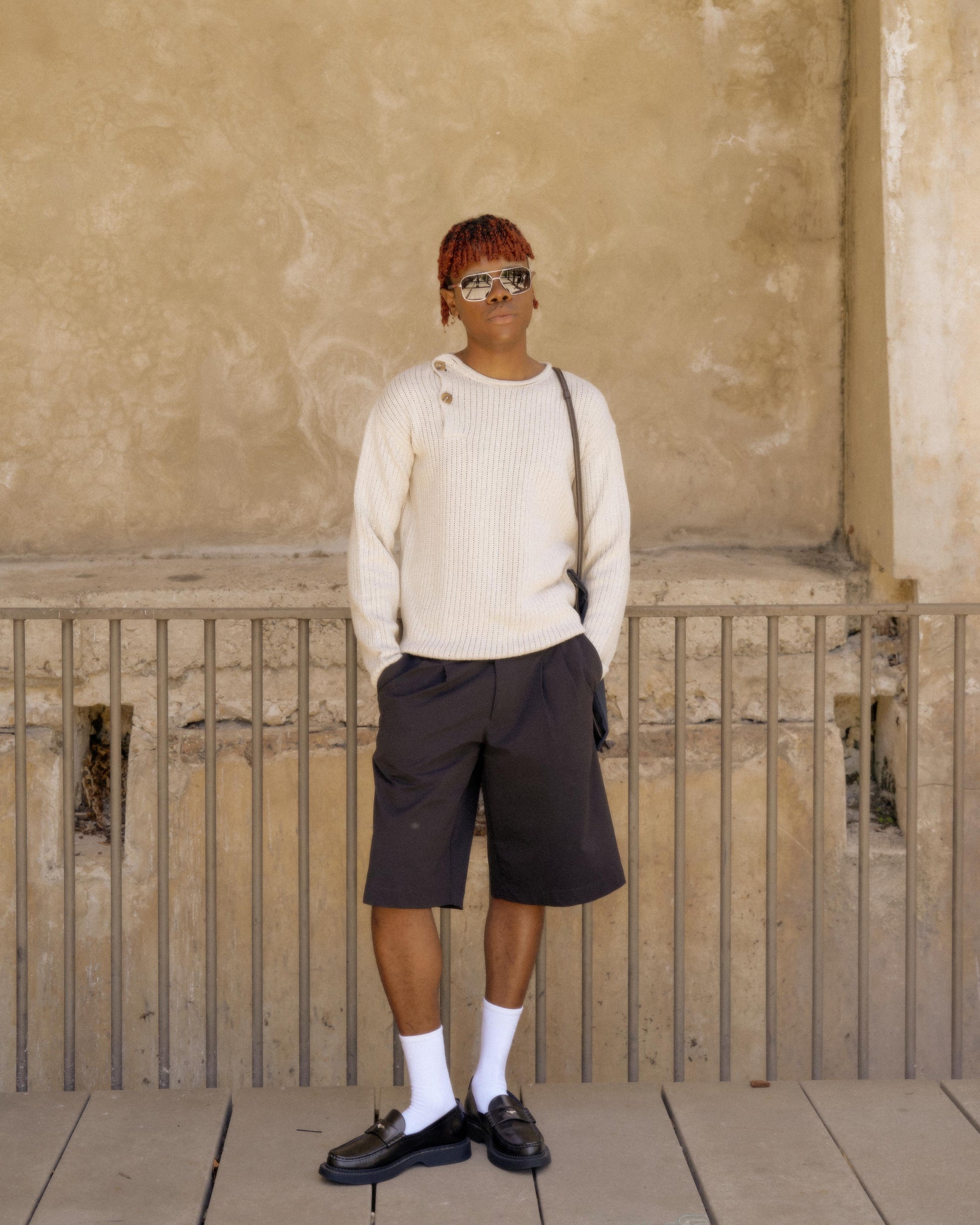 A person standing with one hand in a pocket, wearing a beige knitted Hermès pullover and black shorts, against a textured background.