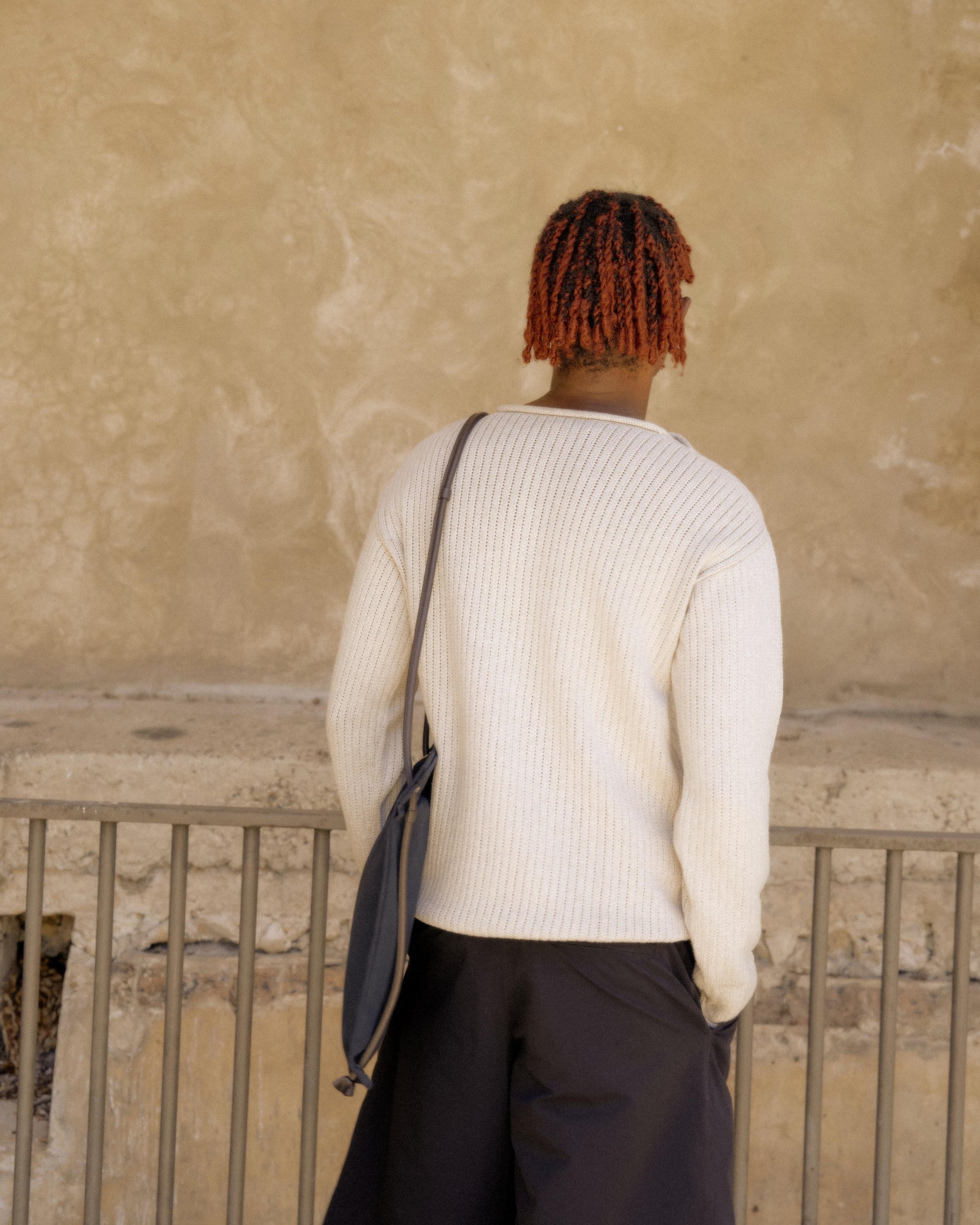 Person with red braids, in a white Hermès sweater and black pants, stands facing a textured beige wall, conveying a calm, introspective mood
