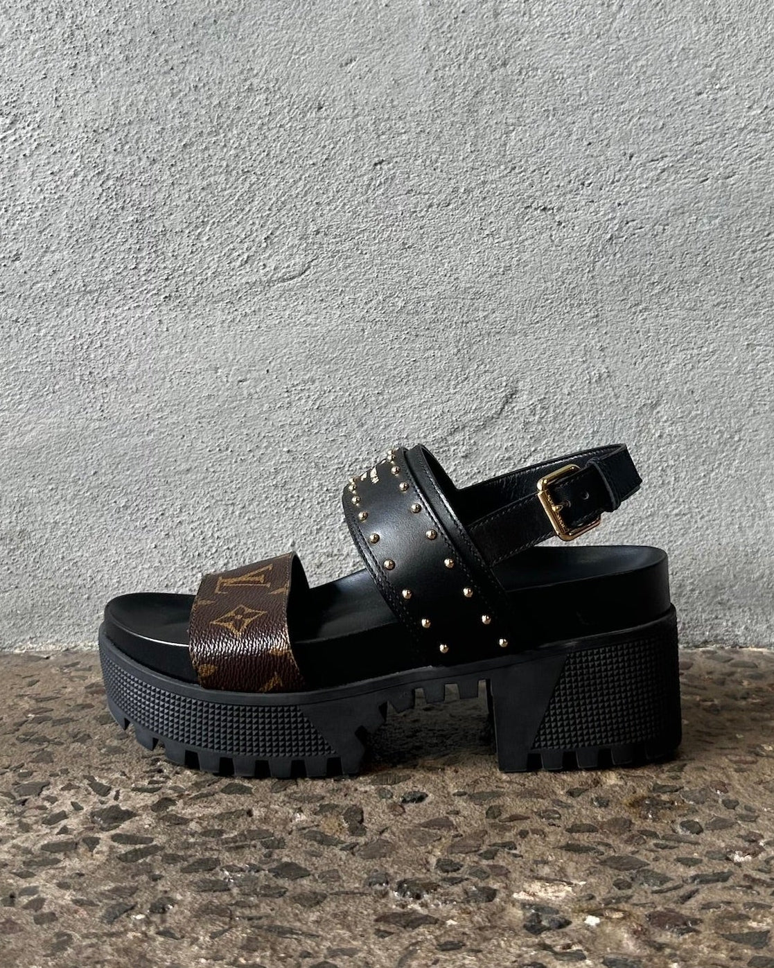 Black sandals with brown strap on a textured surface against a gray wall