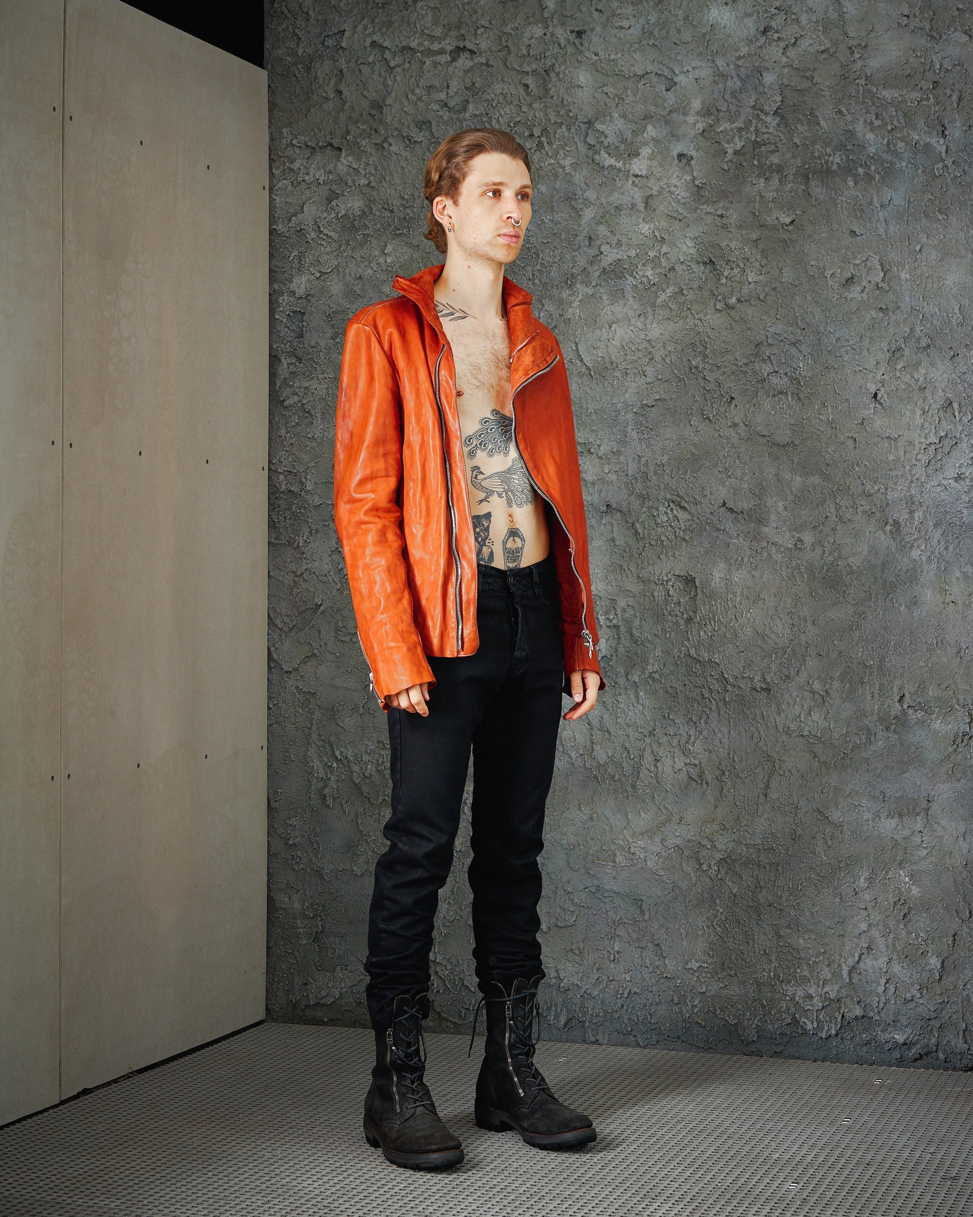 A person stands against a textured grey wall, wearing an open orange Incarnation leather jacket, revealing chest tattoos. They have black pants and boots