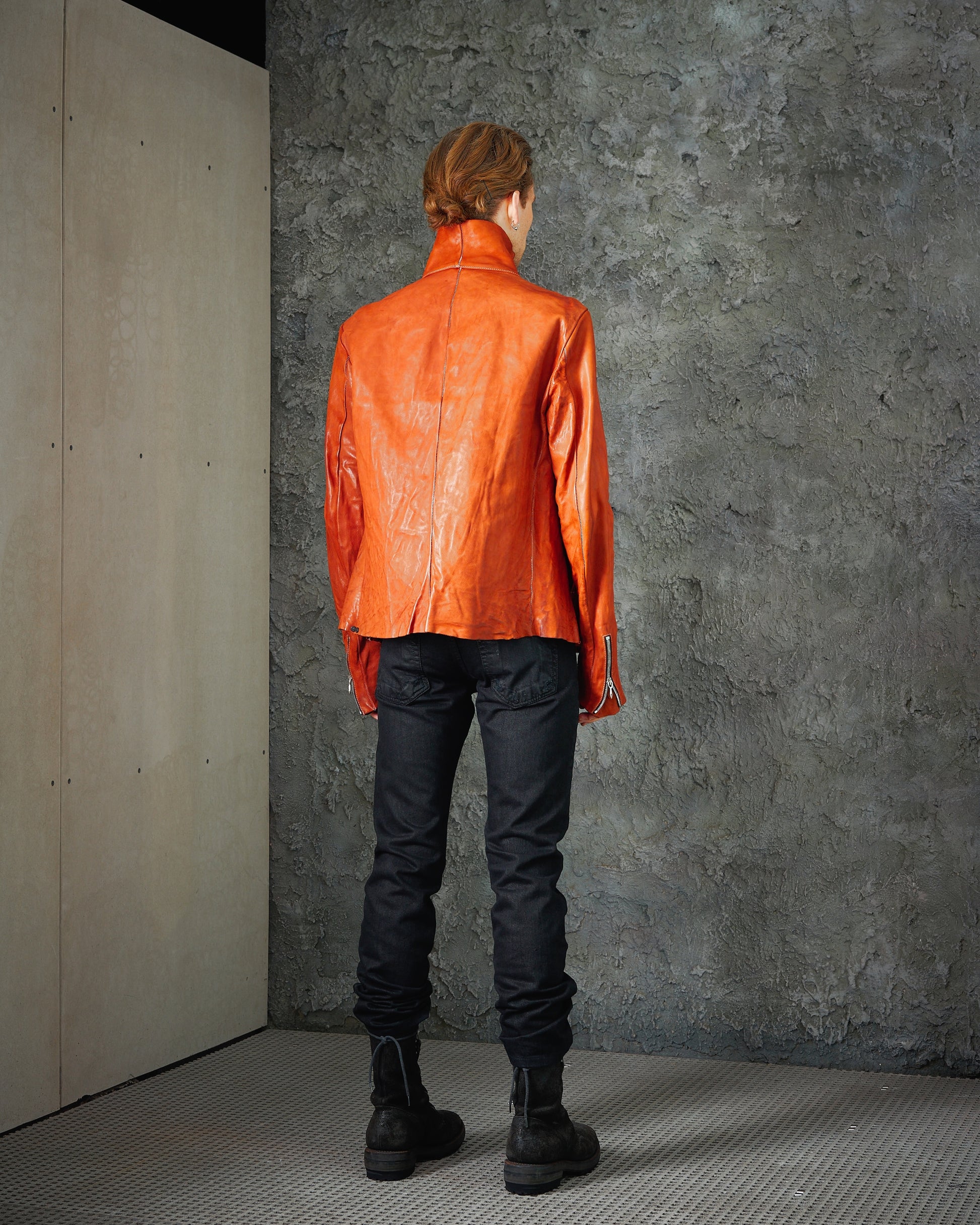 A person stands facing a textured grey wall, wearing an orange leather Incarnation jacket and black jeans. 