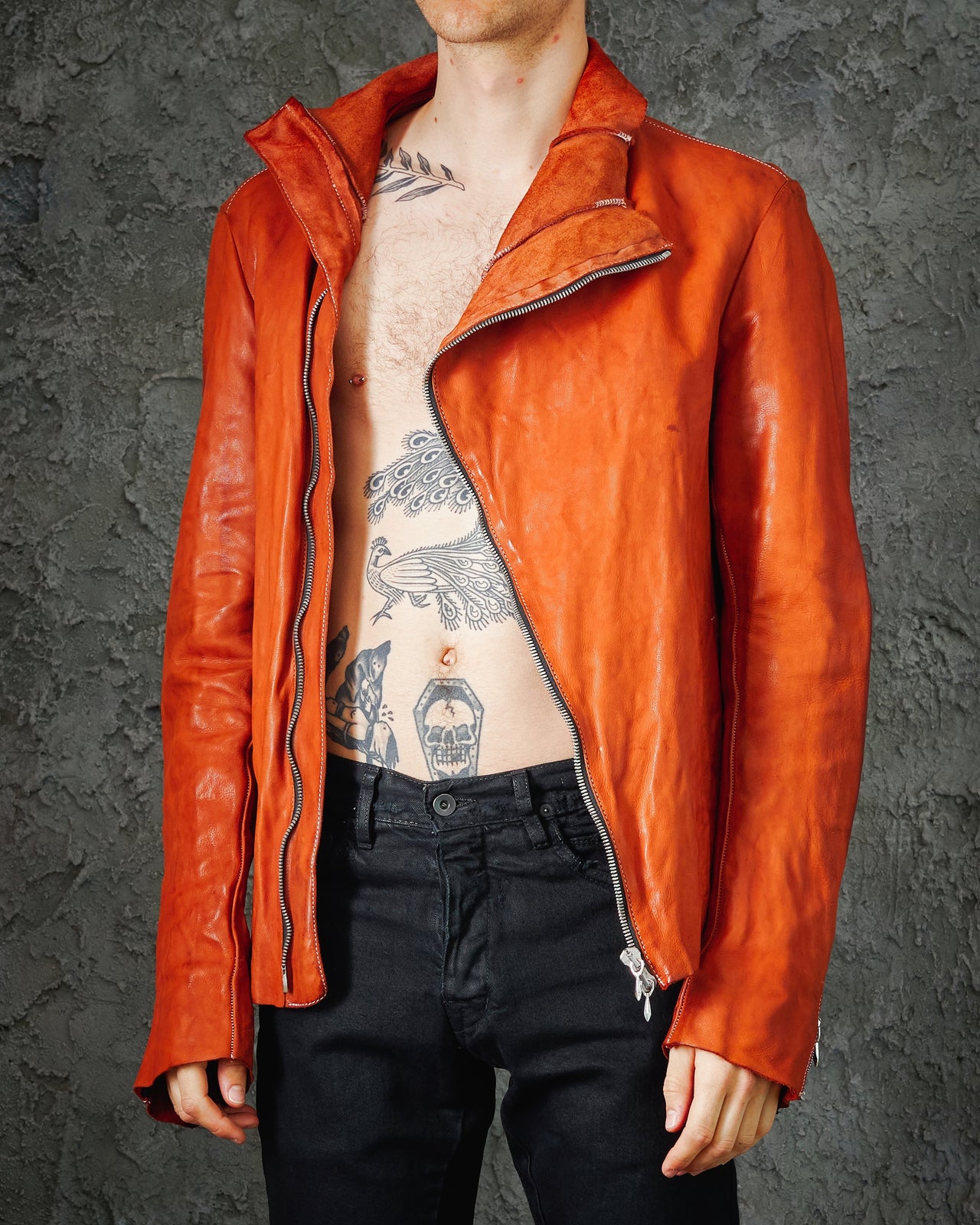 A male model wearing an orange, asymmetrical Incarnation zip leather jacket with a sculptural collar and darted structure, paired with black pants and black boots.