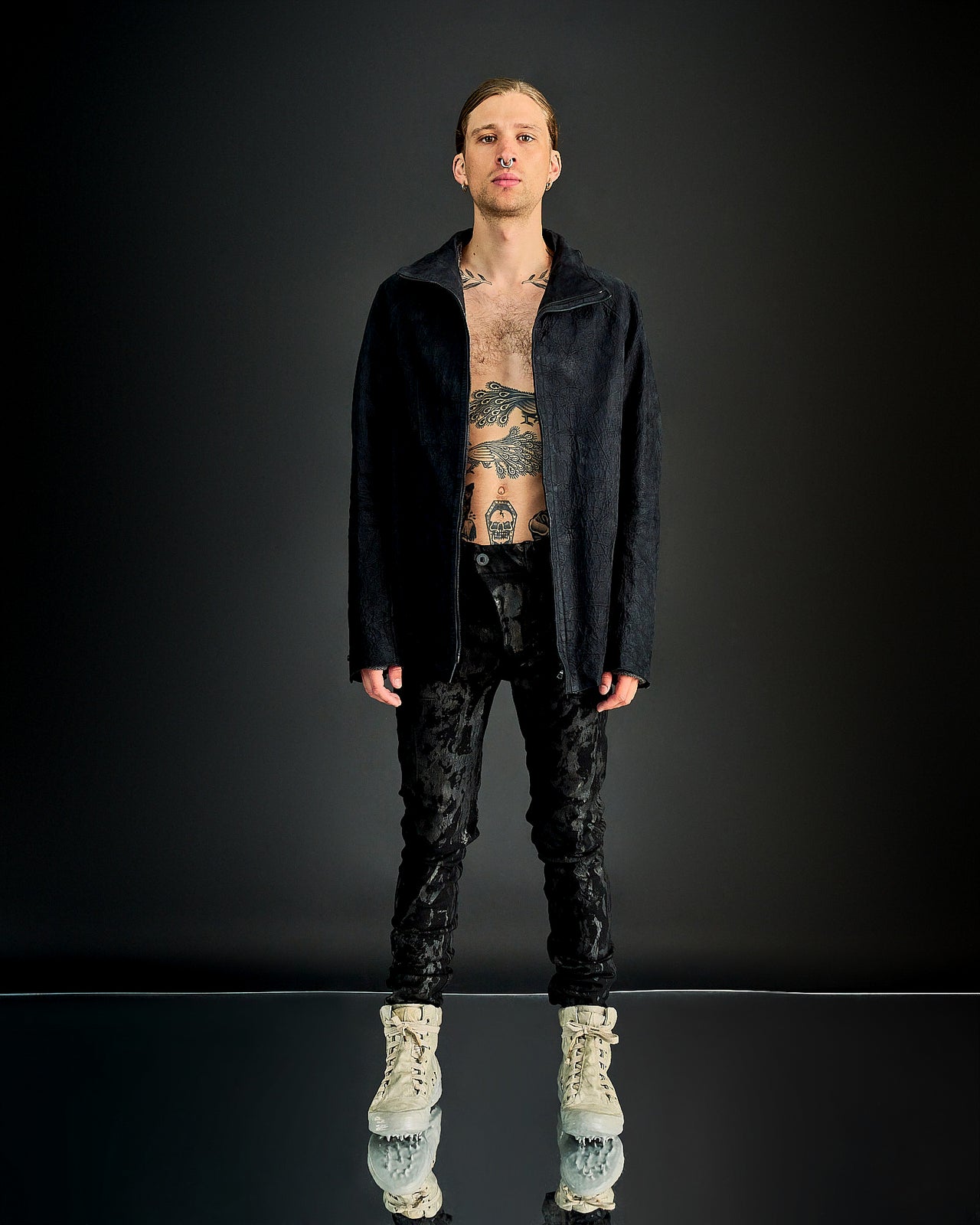 A person standing in a dark setting, wearing a black leather Isaac Sellam jacket with a front zip and textural details, paired with black pants. The person has tattoos and is accessorised with a beige hat and shoes.