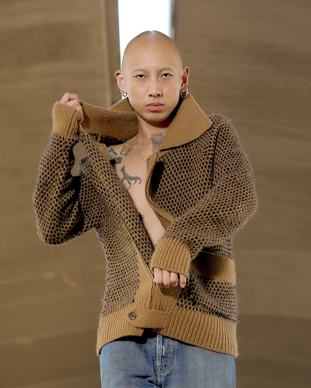 A person with a shaved head poses confidently in a textured brown Issey Miyake cardigan, slightly open to reveal a tattooed chest. They wear blue jeans and hoop earrings.