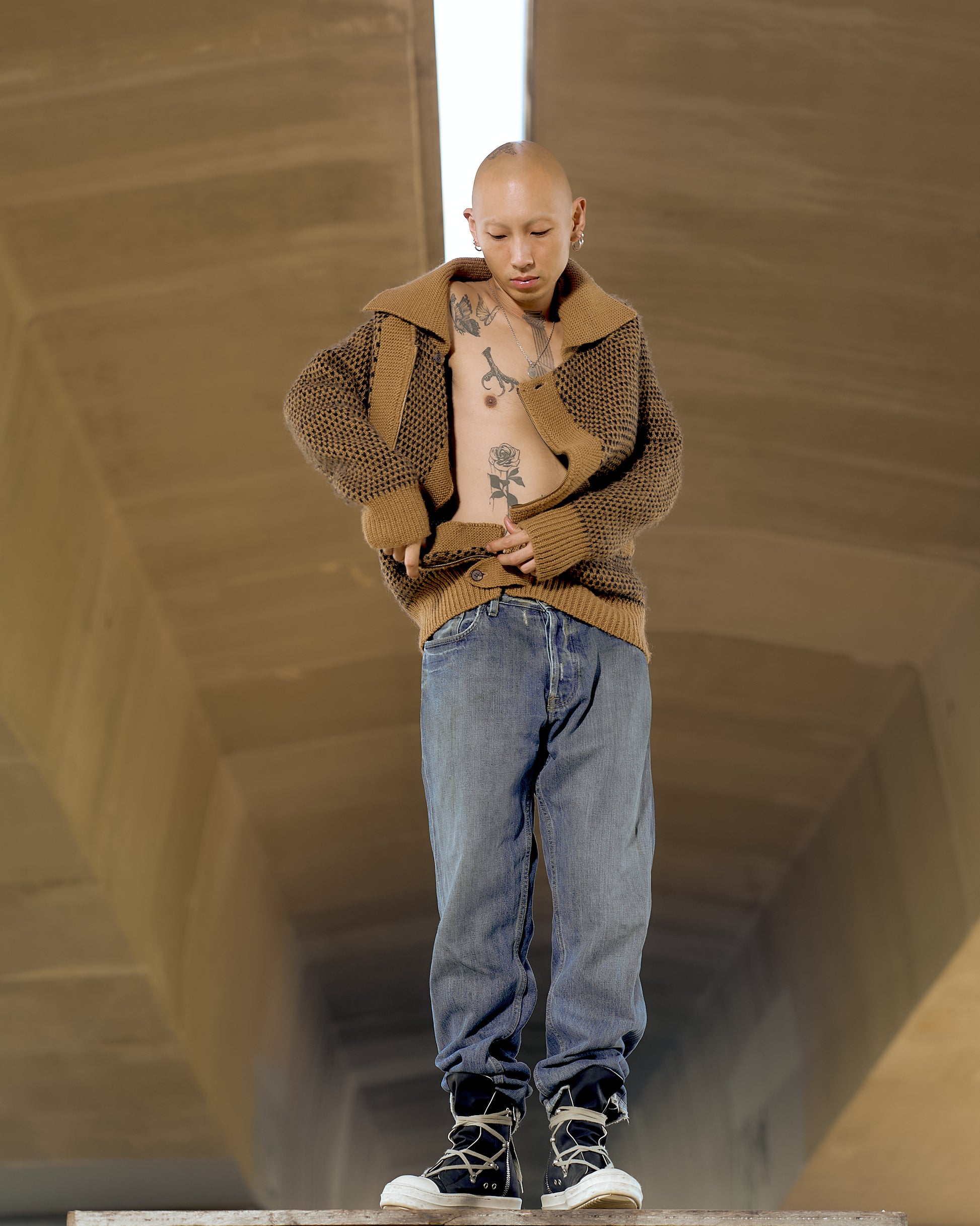 A person with a shaved head stands confidently, displaying tattoos on their torso. They wear a knitted brown Issey Miyake sweater, blue jeans, and black sneakers. T