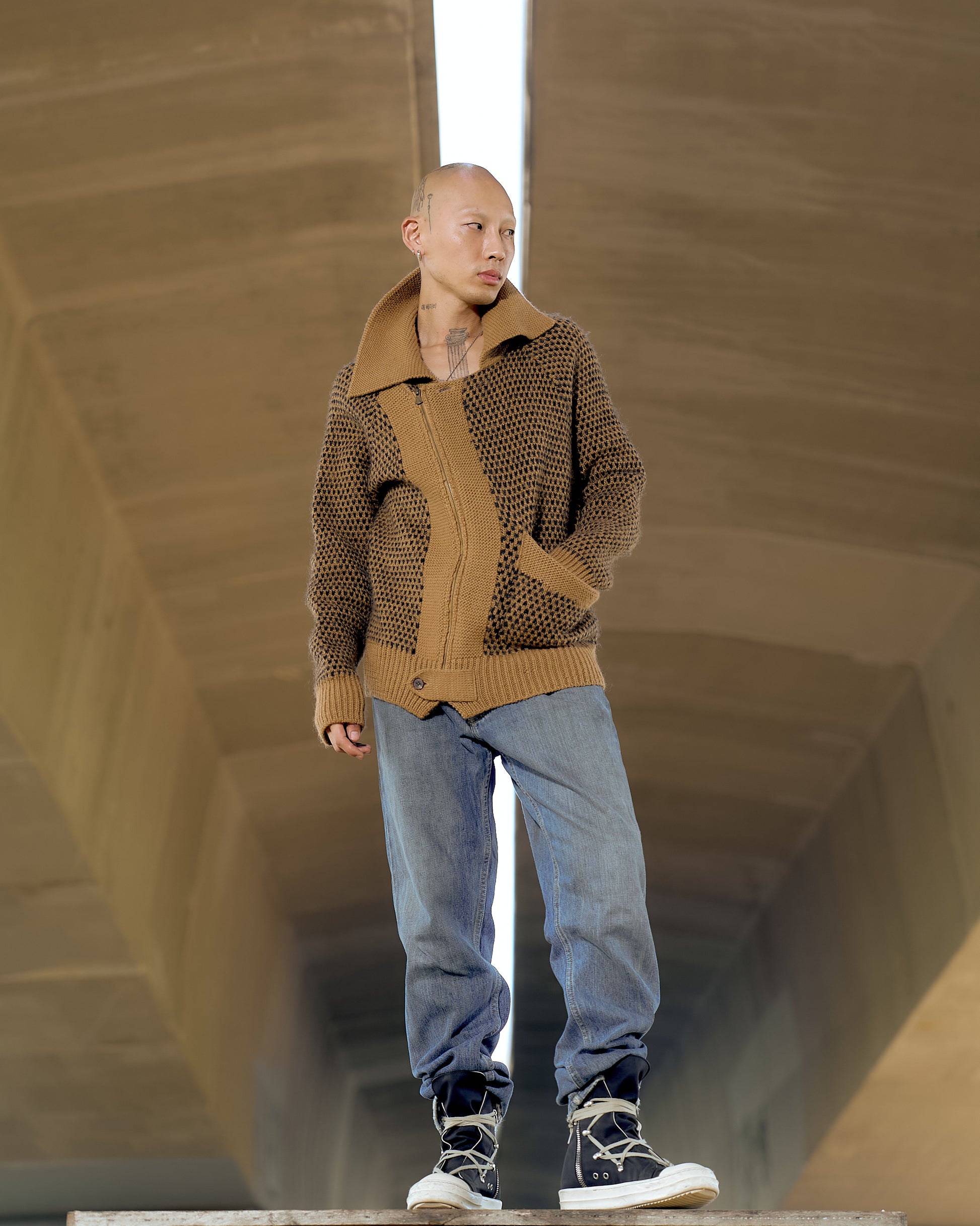 A person wearing a brown collared Issey Miyake knit zip cardigan with wide pattern and raglan sleeves, paired with blue jeans and sneakers, standing under a bridge.