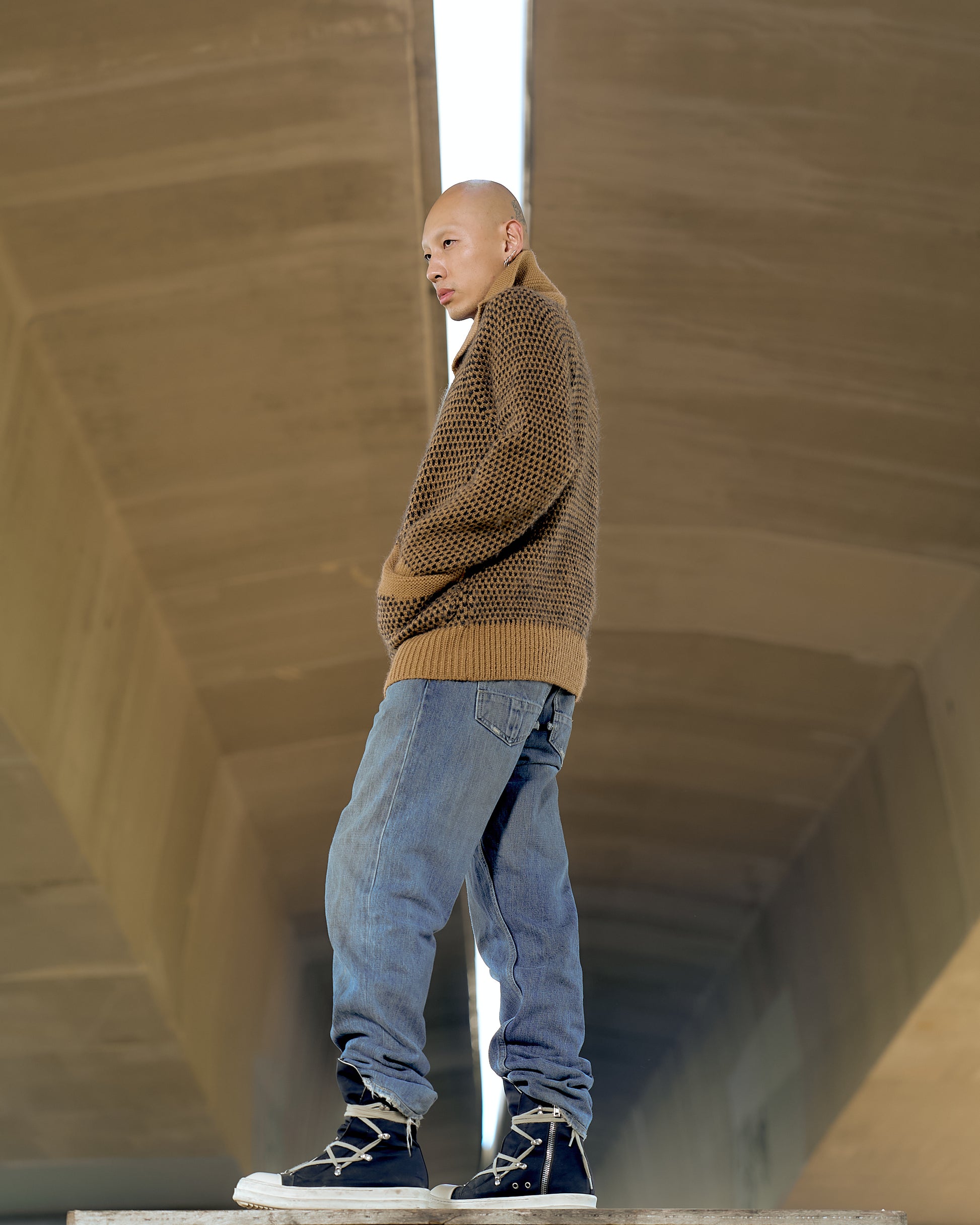 A person stands confidently on a concrete surface, wearing a patterned brown Issey Miyake sweater, blue jeans, and high-top sneakers. The setting is minimal and modern.