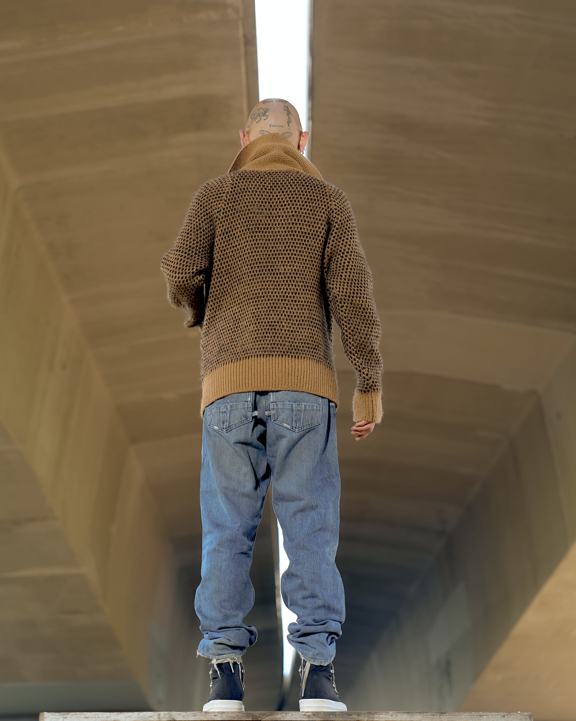 A person in a brown Issey Miyake sweater and blue jeans stands on a concrete surface, seen from behind. The setting is a large tunnel, creating a sense of isolation.