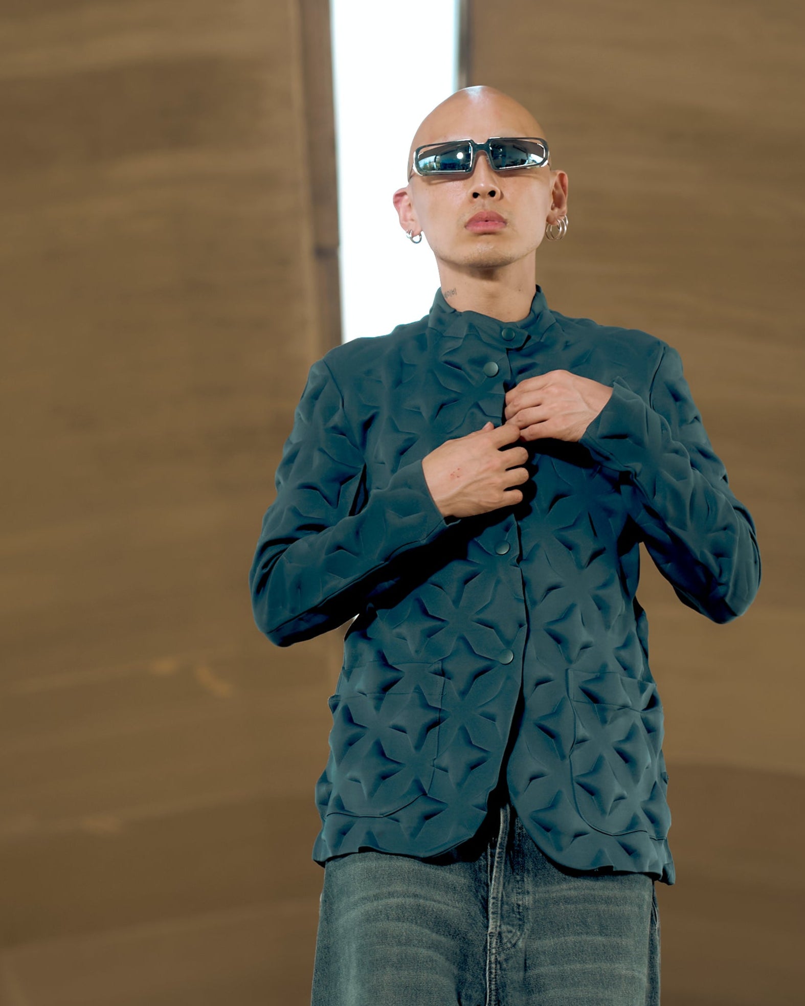 Person in dark sunglasses and a textured dark Issey Miyake jacket stands confidently under a glowing overhead light