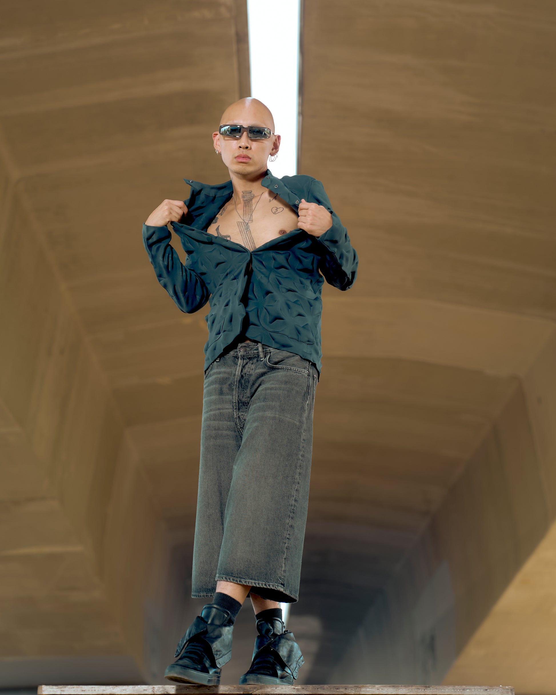 Person in a blue textured Issey Miyake jacket, revealing a tattooed chest, wears oversized sunglasses and wide-leg jeans. They exude confidence and edgy style.