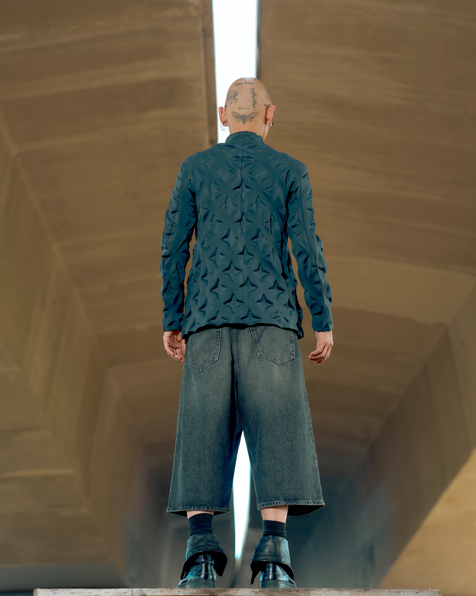 Bald person with head tattoos facing away, wearing a textured Issey Miyake jacket, baggy denim shorts, and black shoes, standing under a concrete structure.