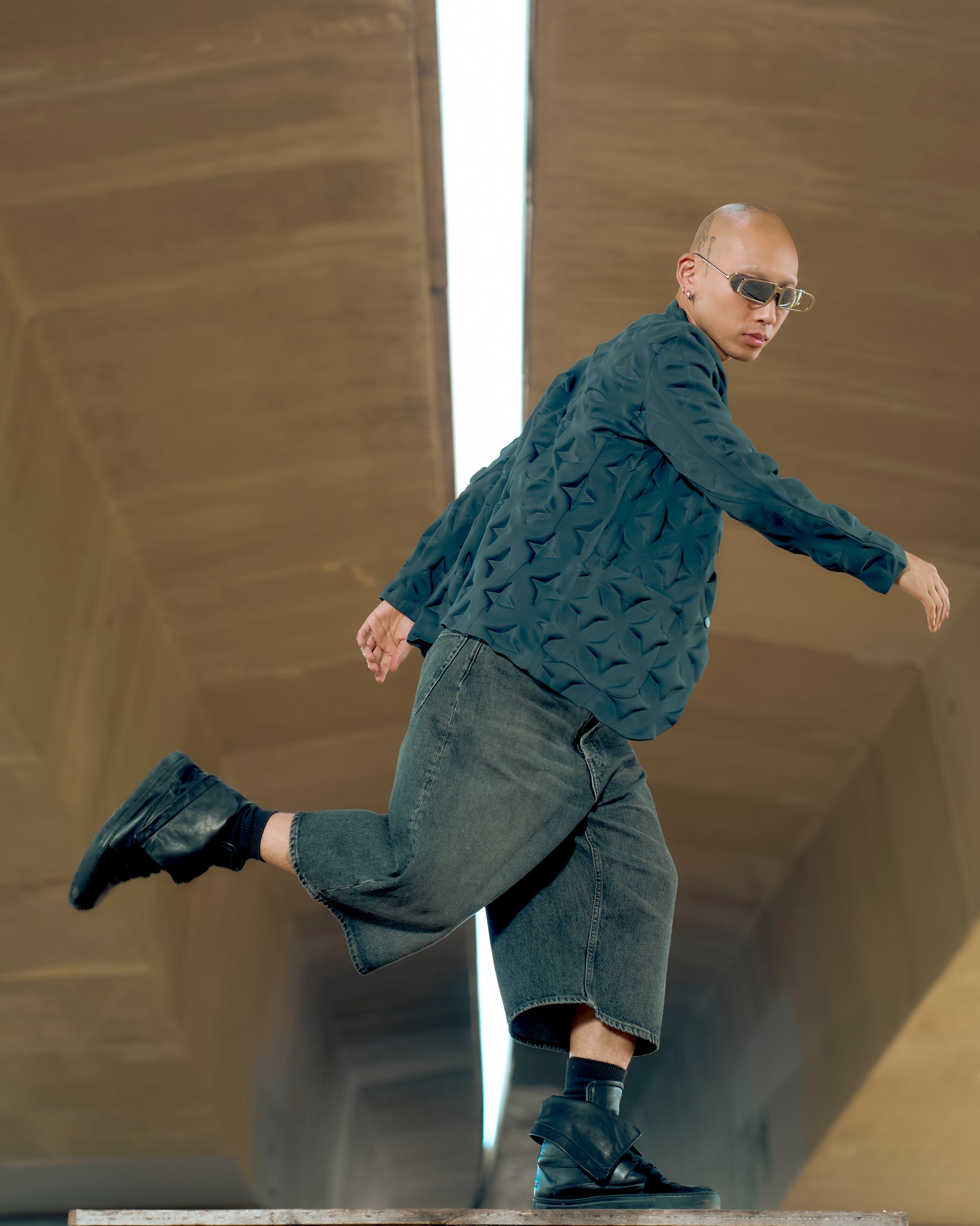 A person in stylish sunglasses and textured Issey Miyake jacket jumps energetically under an overpass. They wear baggy shorts and black high-top sneakers. 