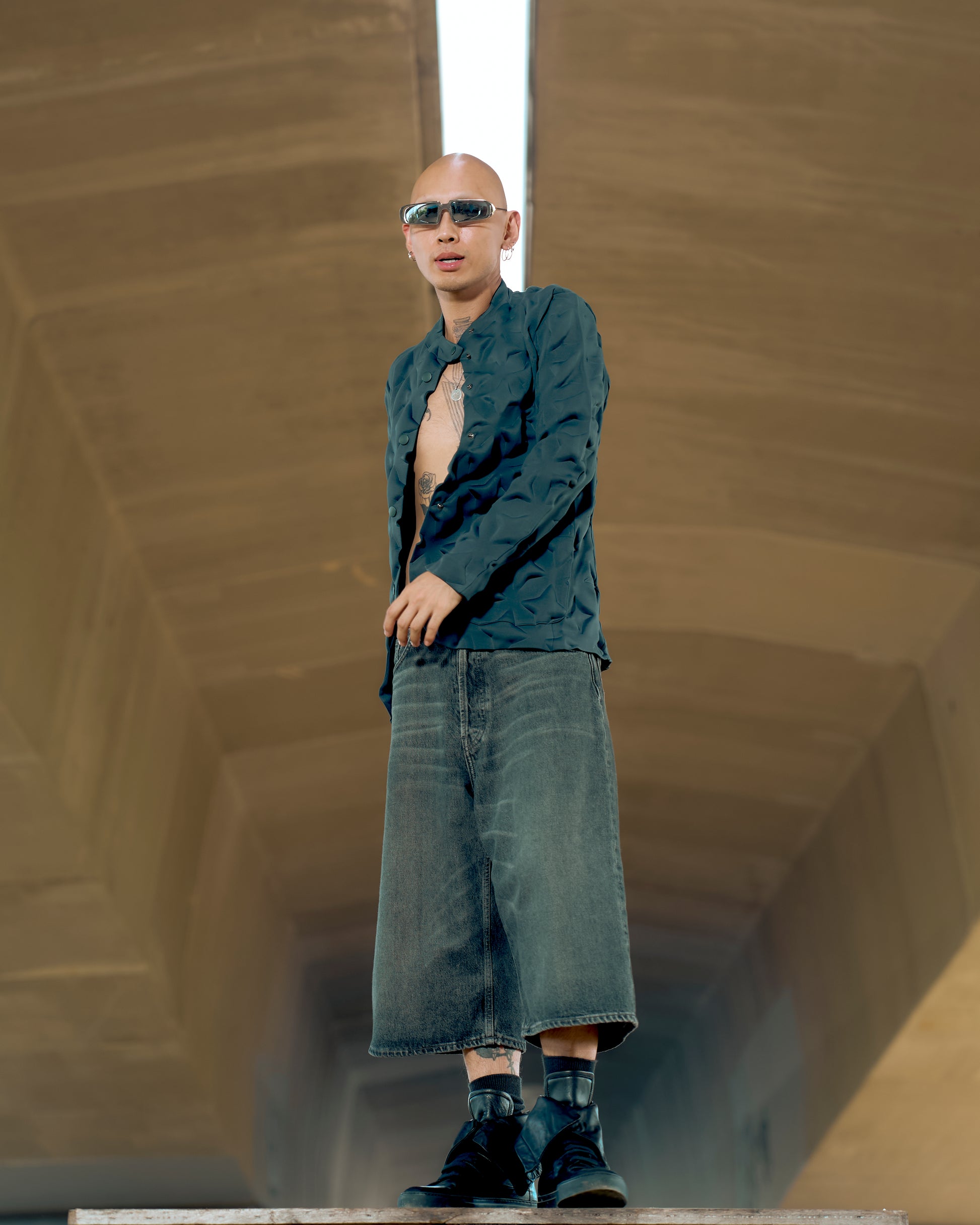 A person in sunglasses and a textured Issey Miyake jacket stands confidently in a tunnel. Wearing oversized denim shorts and boots, they exude a modern, edgy style.
