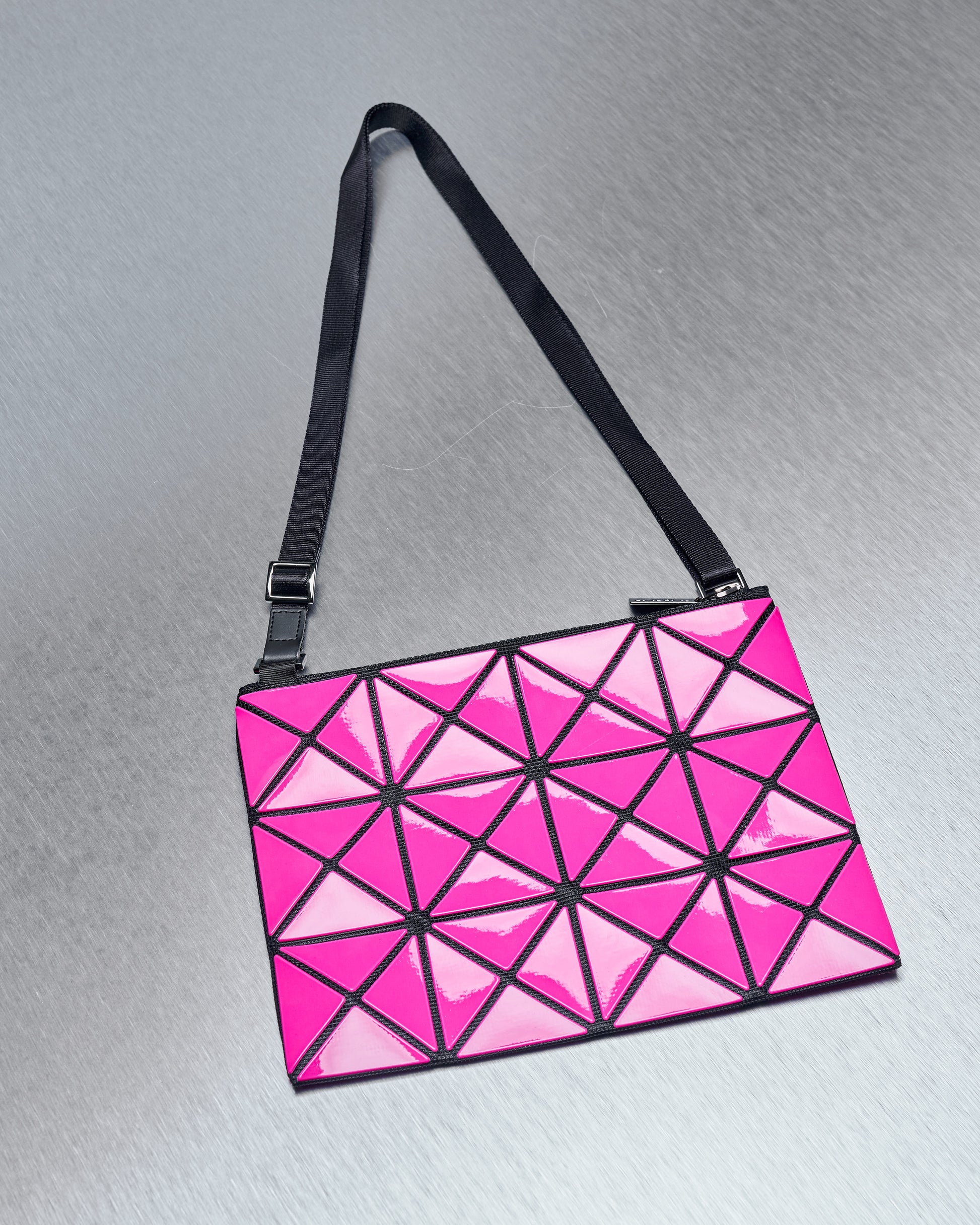 Pink geometric Issey Miyake Bao Bao bag with triangle patterns and a black adjustable strap on a silver metallic surface, conveying a modern, stylish vibe.
