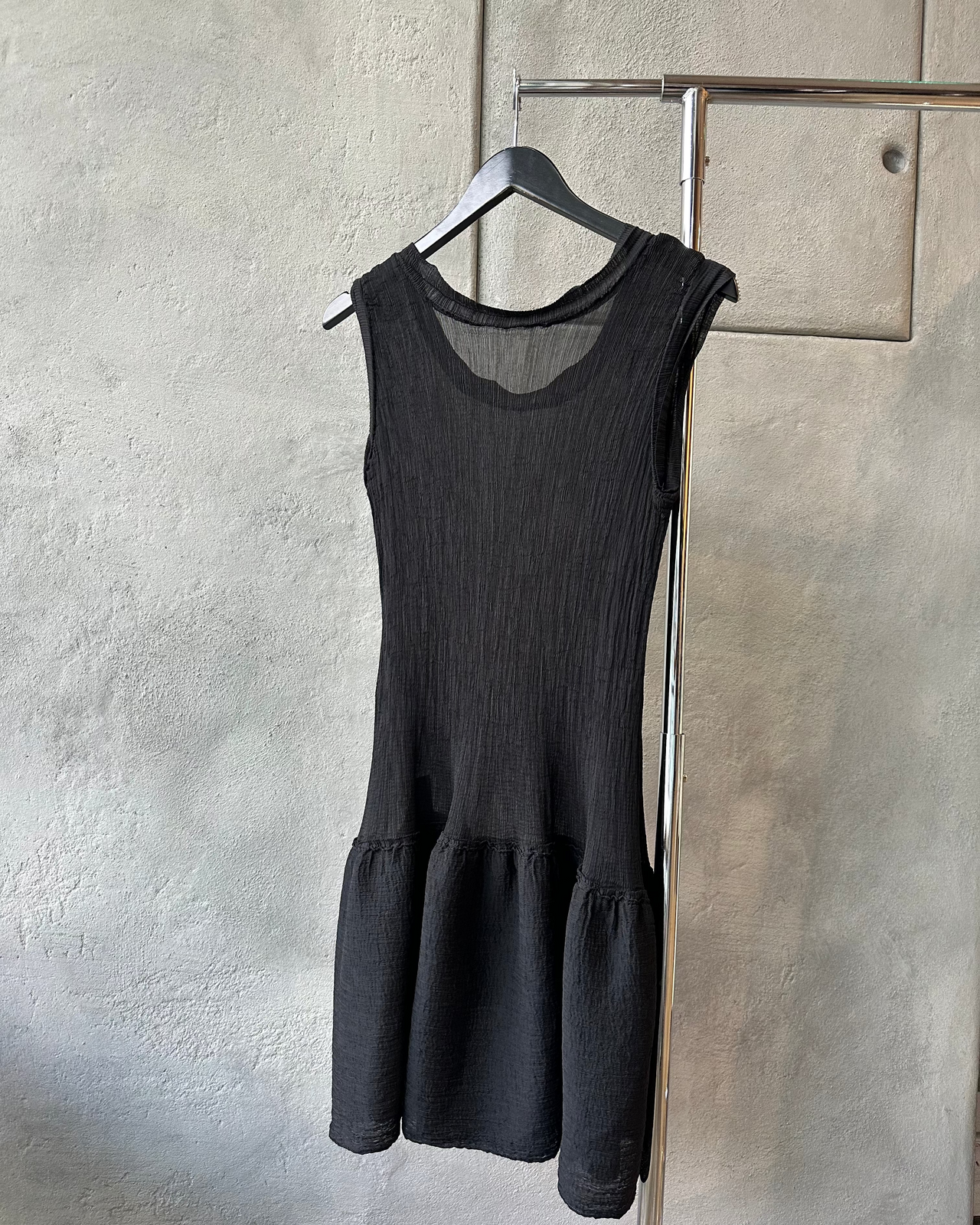 Black sleeveless Issey Miyake dress hanging on a rack against a textured wall.