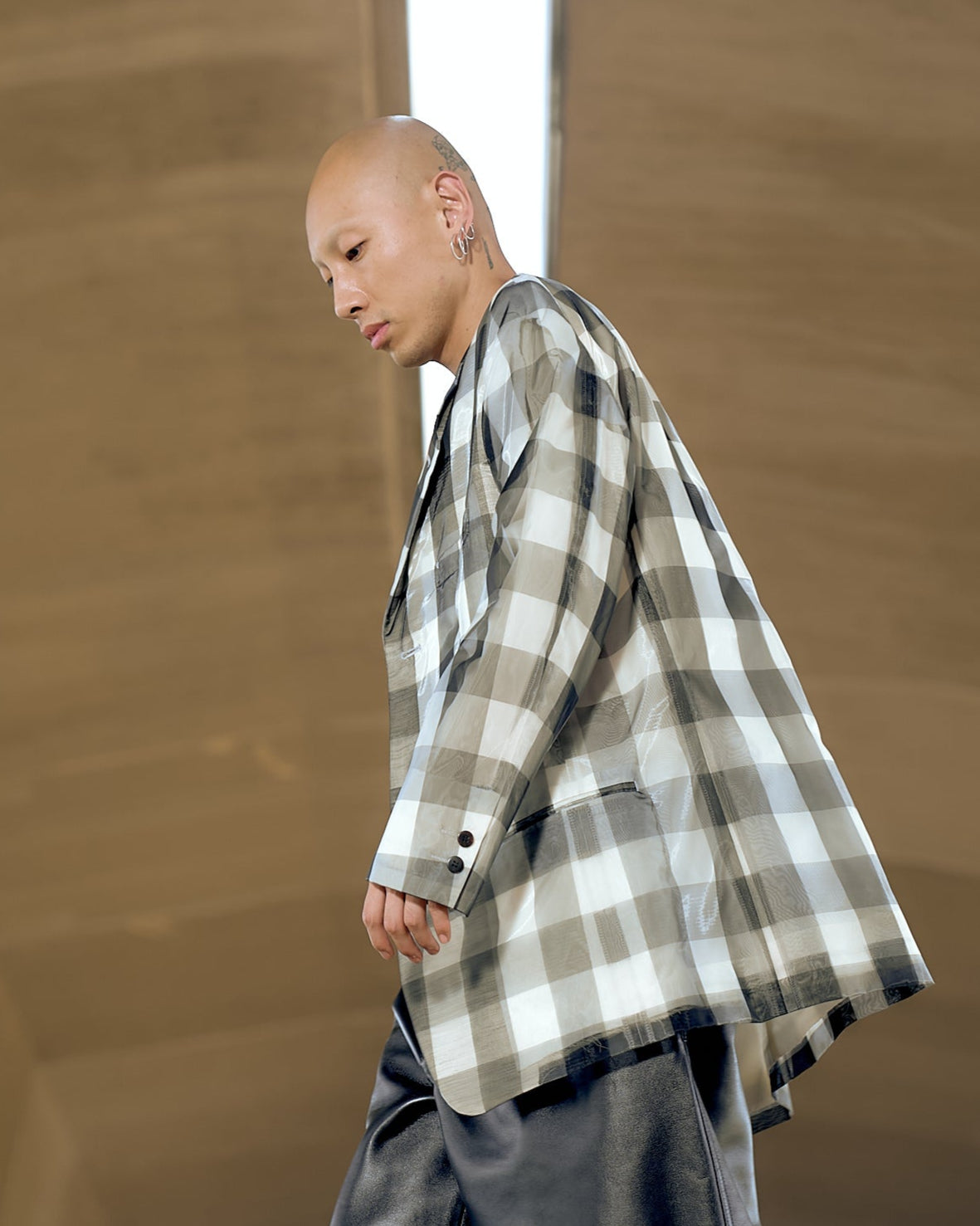 A bald individual in a large plaid Issey Miyake blazer and dark pants looks downward with a focused expression. The background is a neutral, blurred brown.