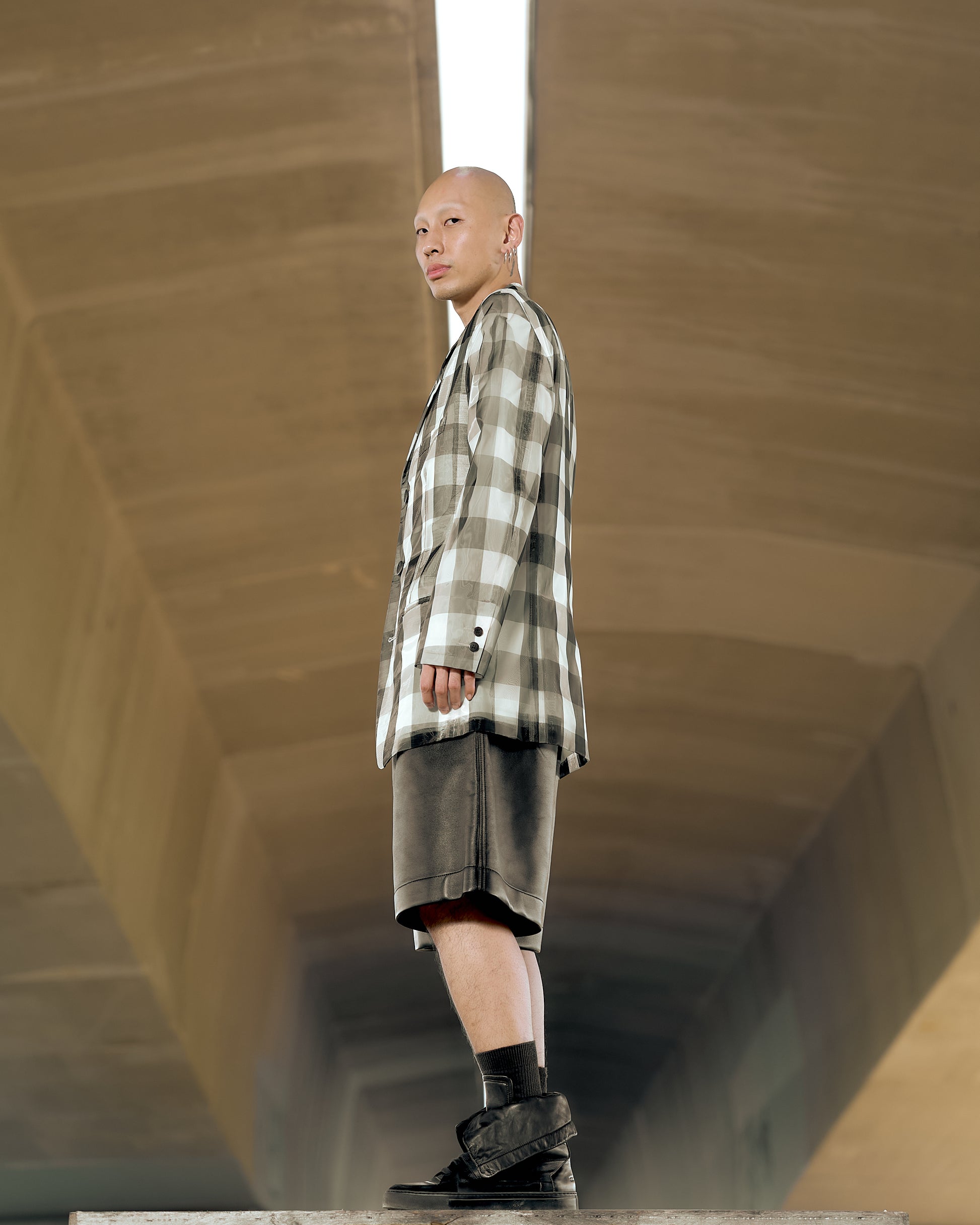 A bald individual in a large plaid Issey Miyake blazer and dark pants looks downward with a focused expression. The background is a neutral, blurred brown.