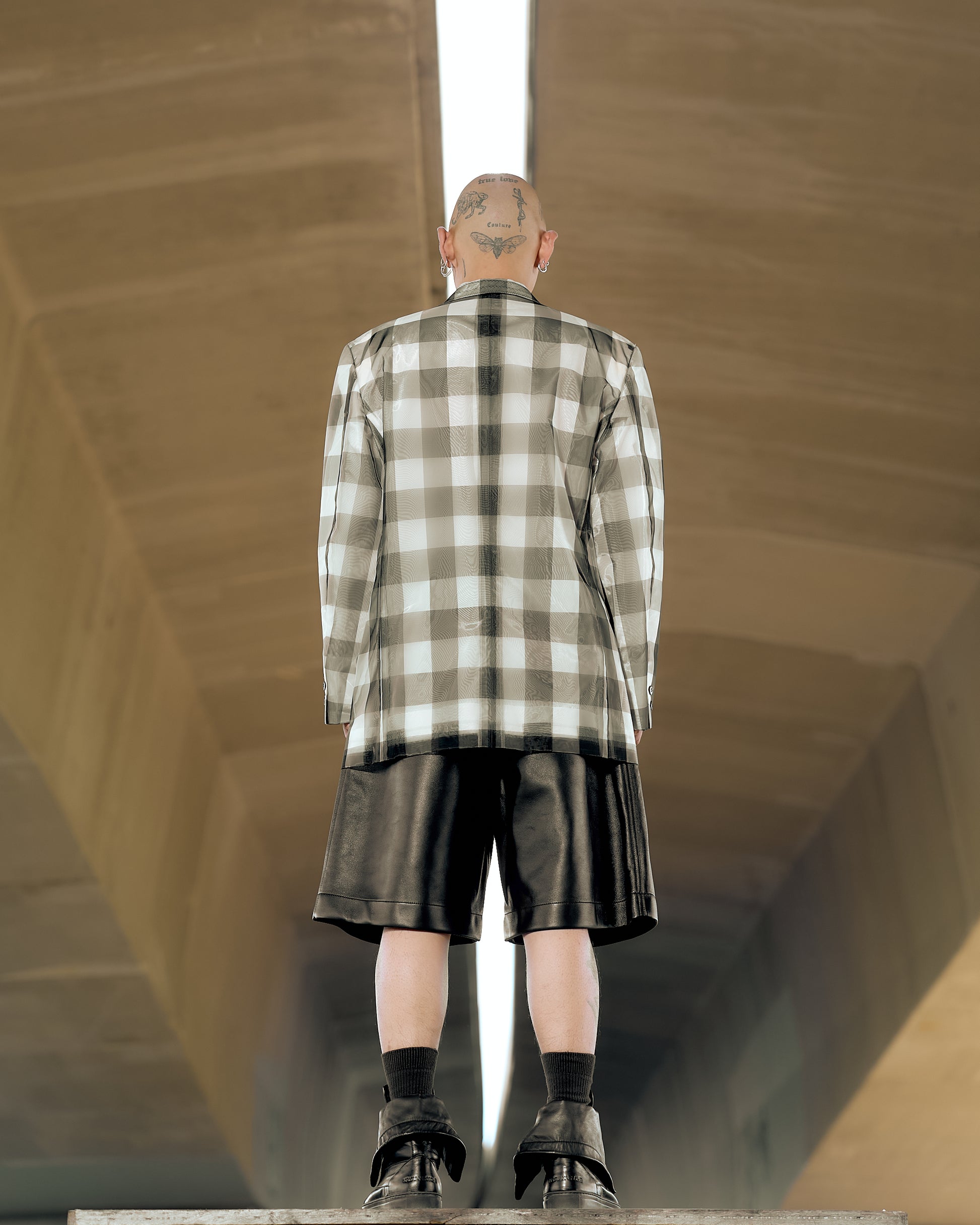 Person stands under a bridge, back facing, wearing a plaid Issey Miyake jacket, black shorts, and boots. Tattoos visible on head. Urban, edgy atmosphere.