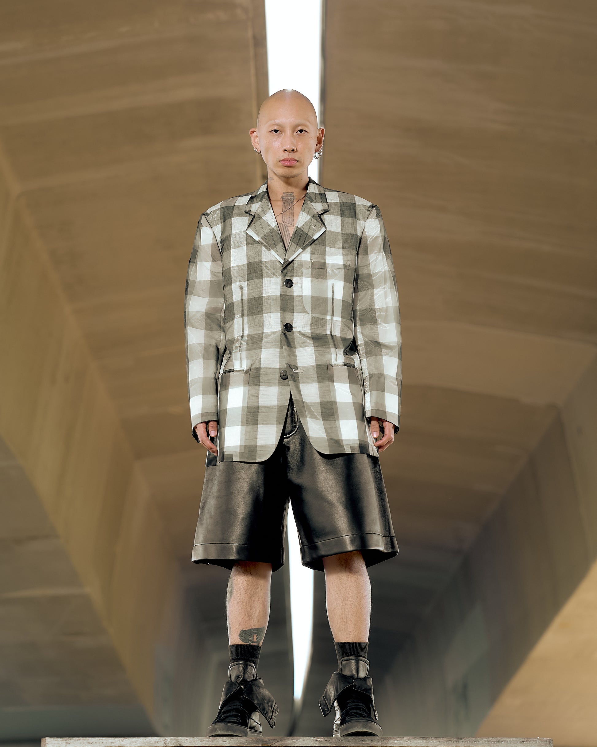 A male model wearing a sheer check Issey Miyake blazer with a peak lapel, three-button closure, and visible endoskeleton structure. The blazer is translucent, revealing the inner structure and materials.