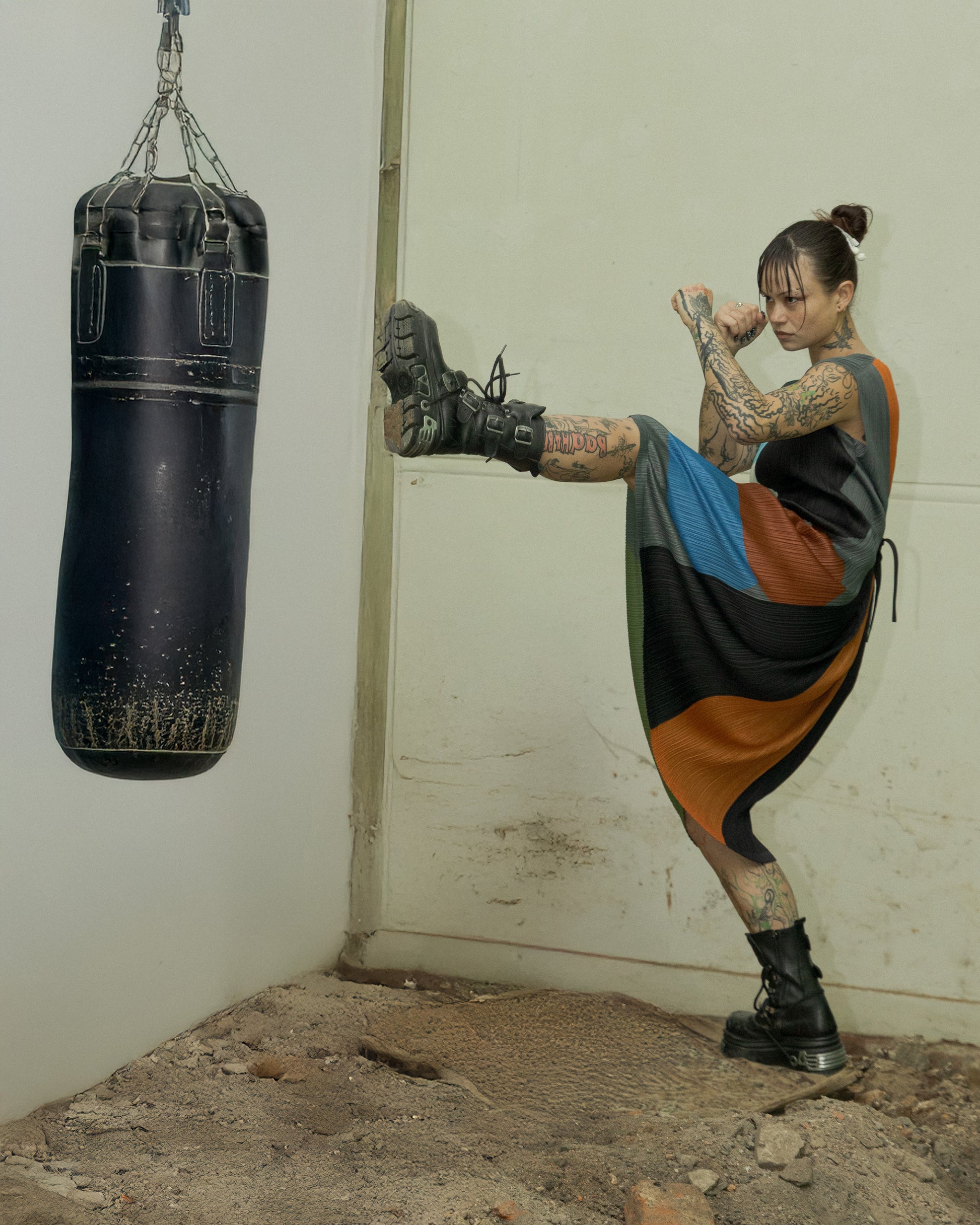 Person in colourful Issey Miyake x Ikko Tanaka outfit performing a kick on a punching bag in a room with a concrete floor.