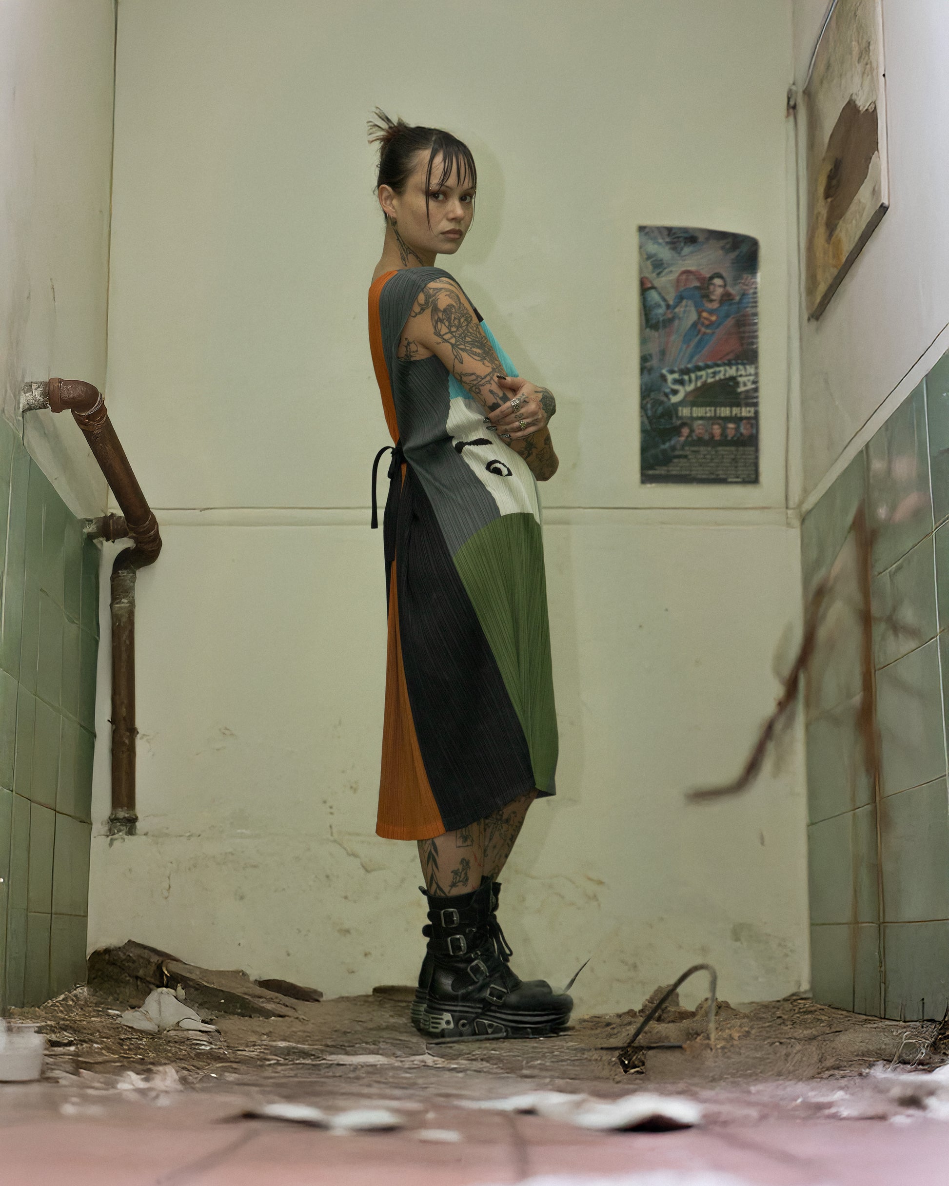 Person with tattoos standing in a dilapidated room with peeling paint and posters on the wall wearing a Issey Miyake dress