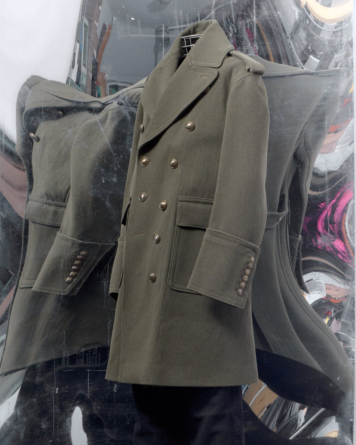 An olive green double-breasted military-style coat displayed on a mannequin, paired with black trousers, set against a reflective mirrored backdrop.
