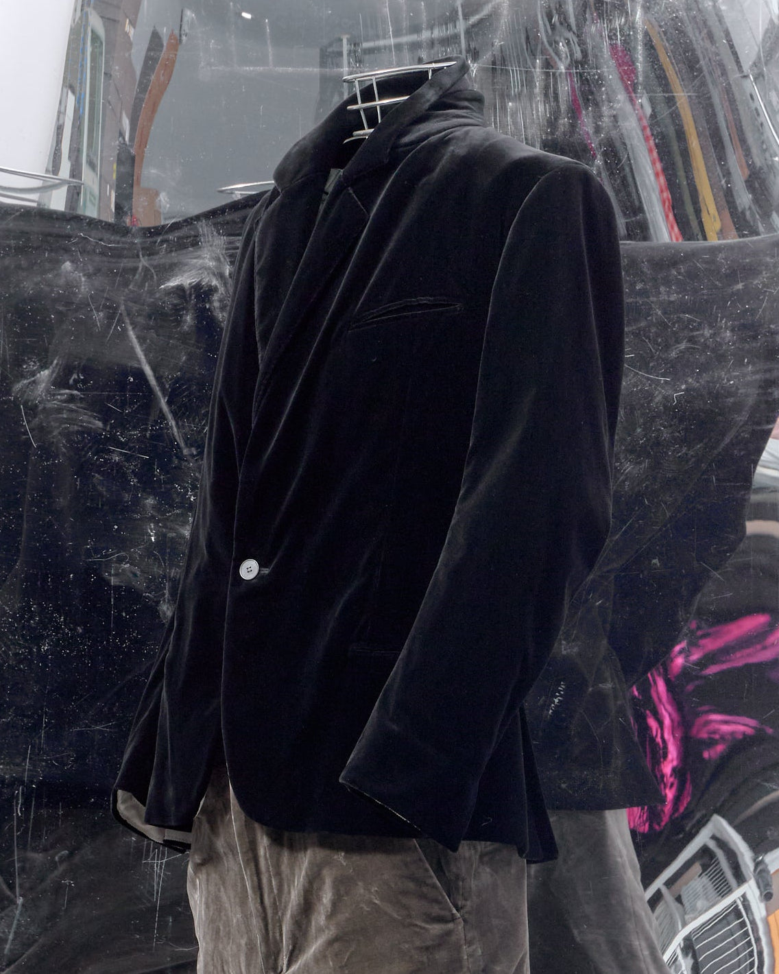 A mannequin wearing a black velvet blazer and brown velvet trousers, displayed in front of a large mirrored wall showing an abstract reflection.