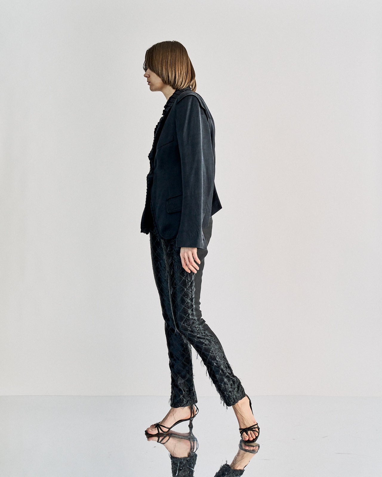 A person wearing black slim-fit JW Anderson trousers with a diamond motif, featuring two side pockets and a straight silhouette, with a zip and button closure.