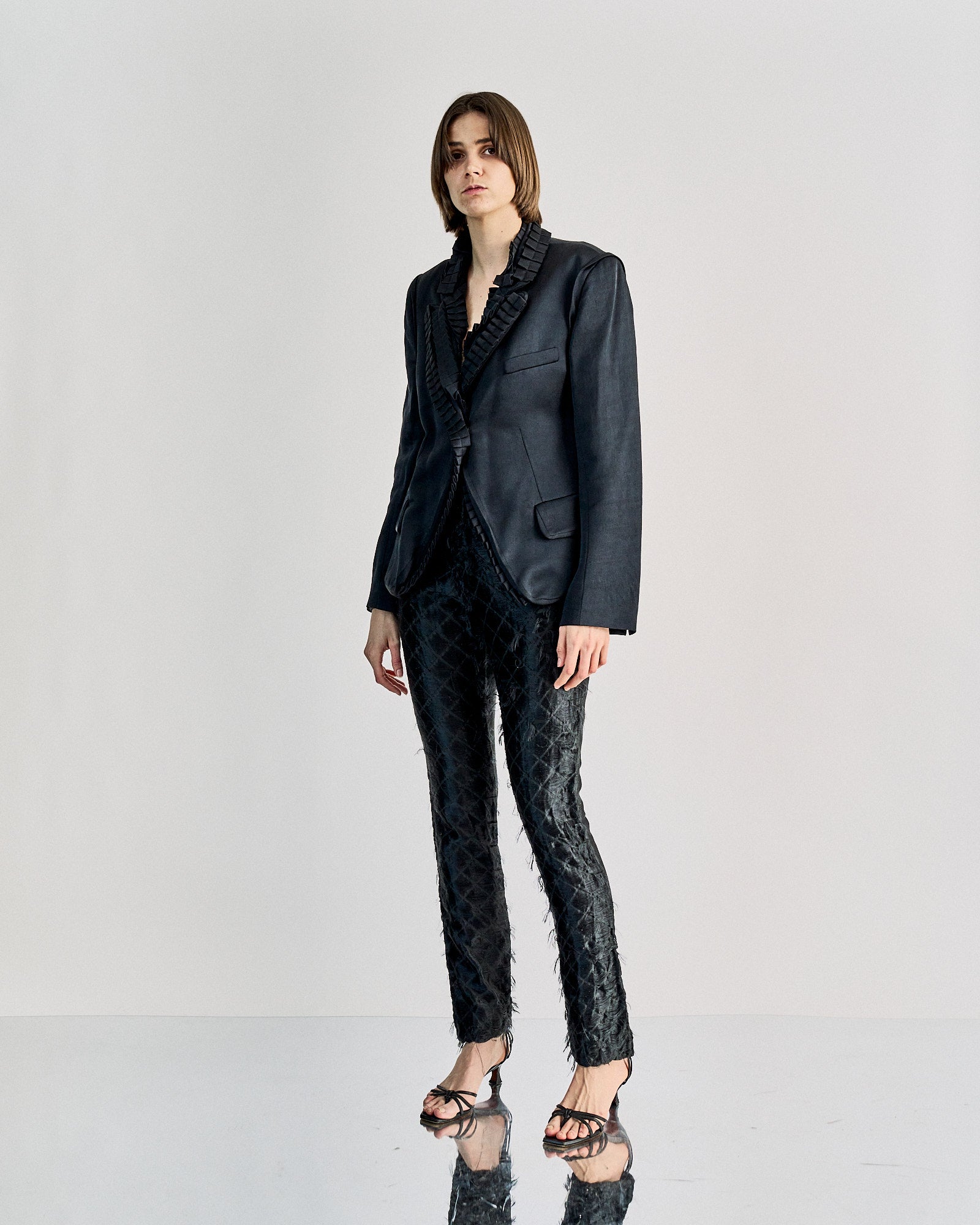 A person stands against a white background, wearing a dark, tailored jacket and textured JW Anderson pants. They project a confident, elegant demeanor.