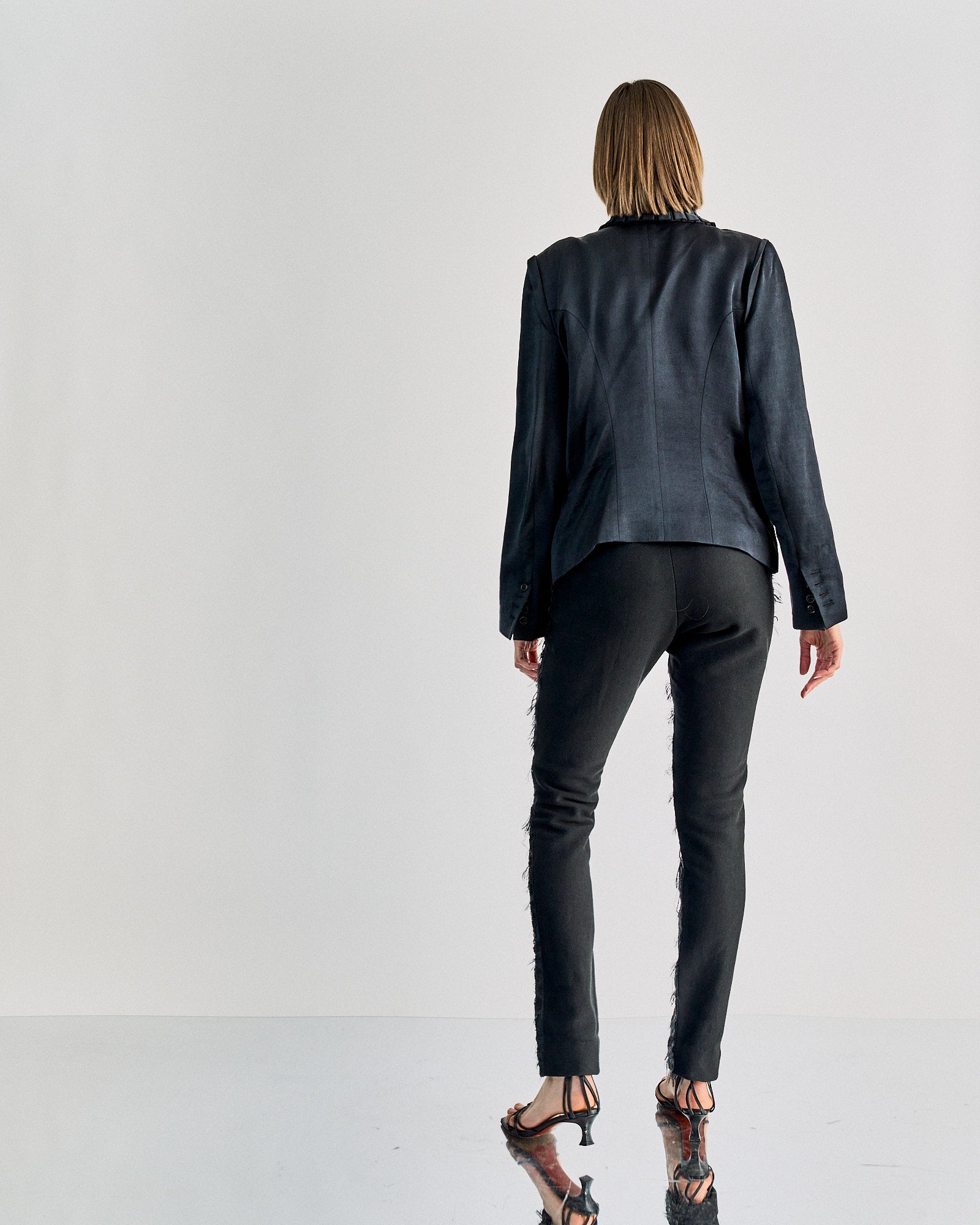A person stands facing away, wearing a fitted black blazer and skinny JW Anderson black pants. The outfit is complemented by strappy black heels. The background is plain white, creating a minimalist and sophisticated tone.