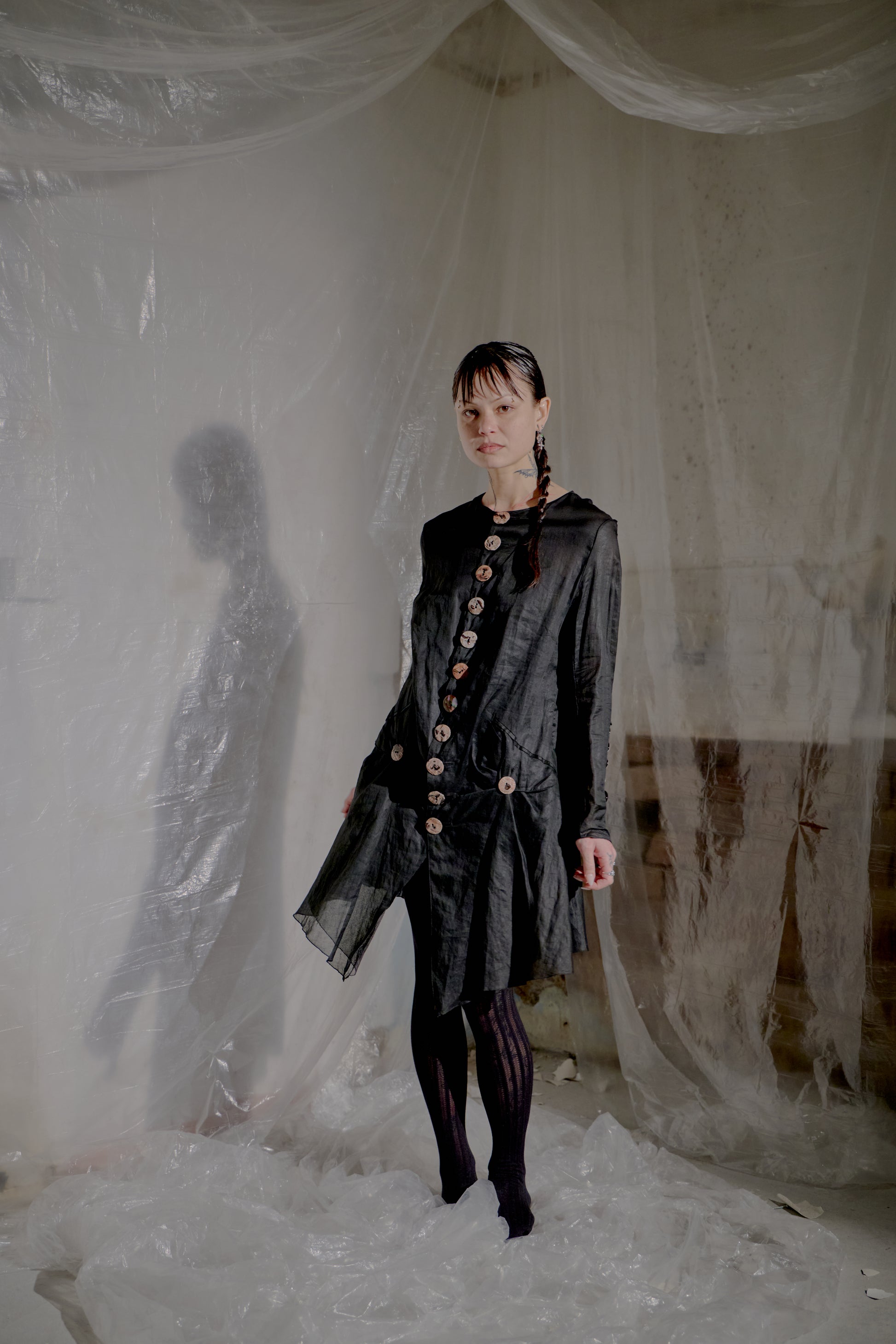 Person wearing a dark Jean Paul Gaultier jacket with buttons in a room with white curtains.