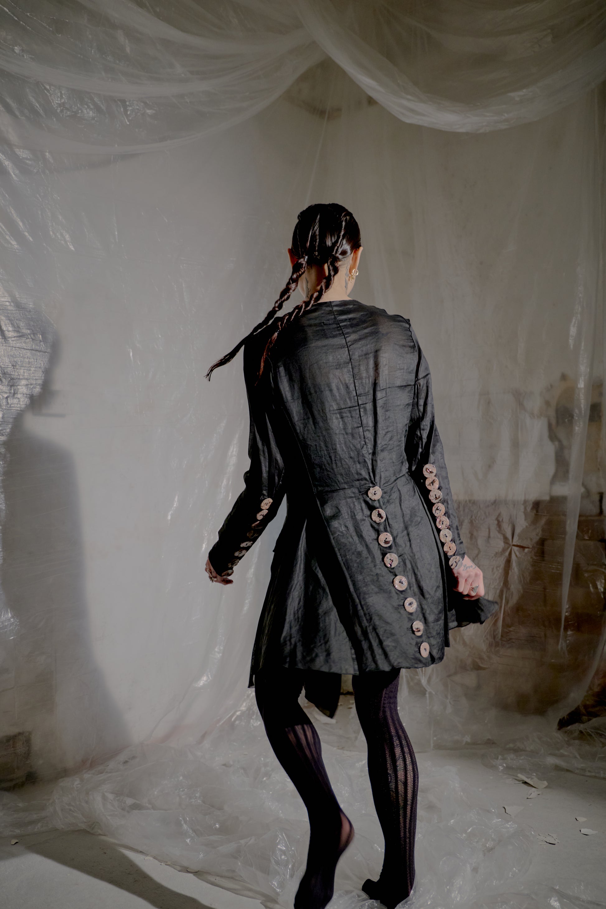 Person wearing a dark Jean Paul Gaultier jacket with decorative buttons in a minimalistic setting.