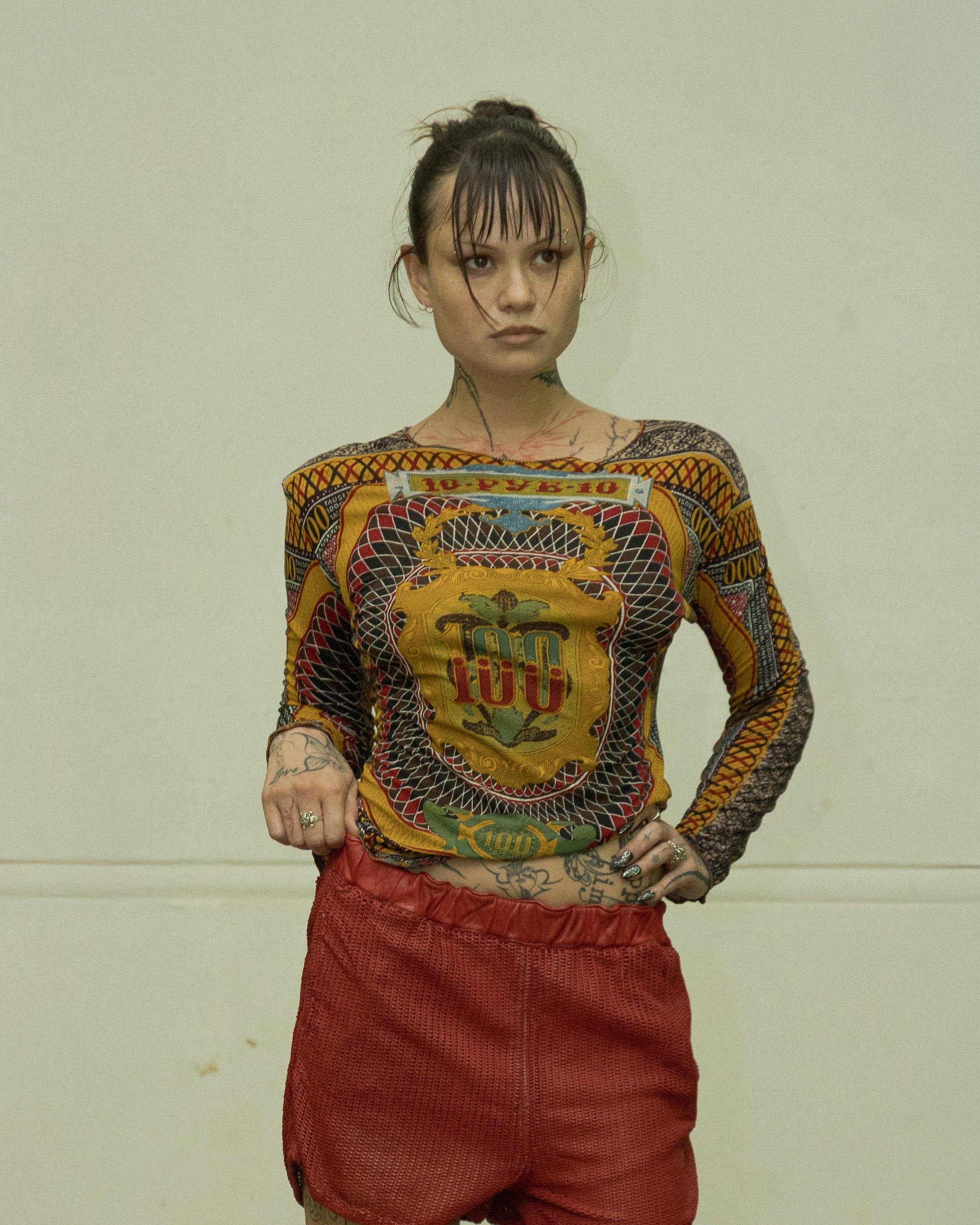 Person wearing a patterned Jean Paul Gaultier top and red shorts standing in a room with a desk and curtains.