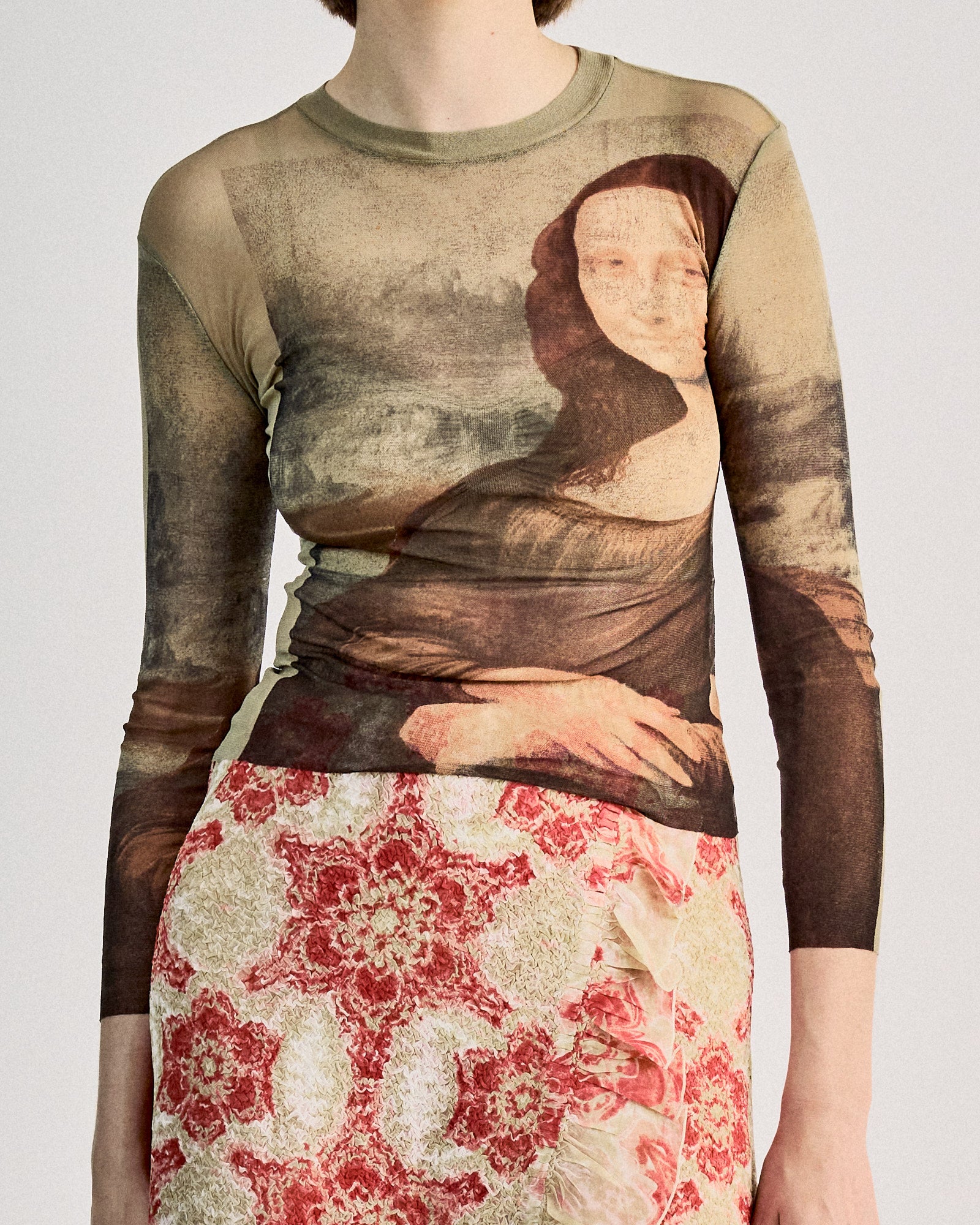 A person wears a sheer, tight-fitting Jean Paul Gaultier top featuring the Mona Lisa print, paired with a skirt with red and white floral patterns, exuding an artistic vibe.