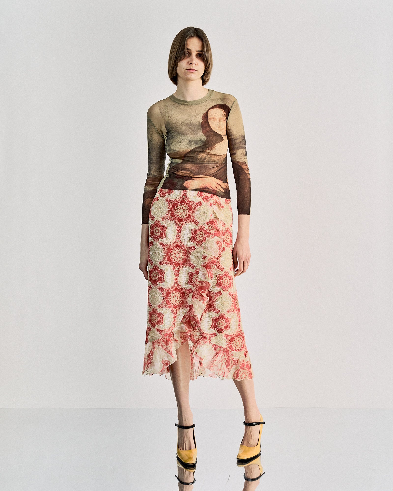 A person stands wearing a long-sleeved Jean Paul Gaultier top printed with the Mona Lisa and a floral-patterned skirt. The setting is minimalist, evoking a modern tone.