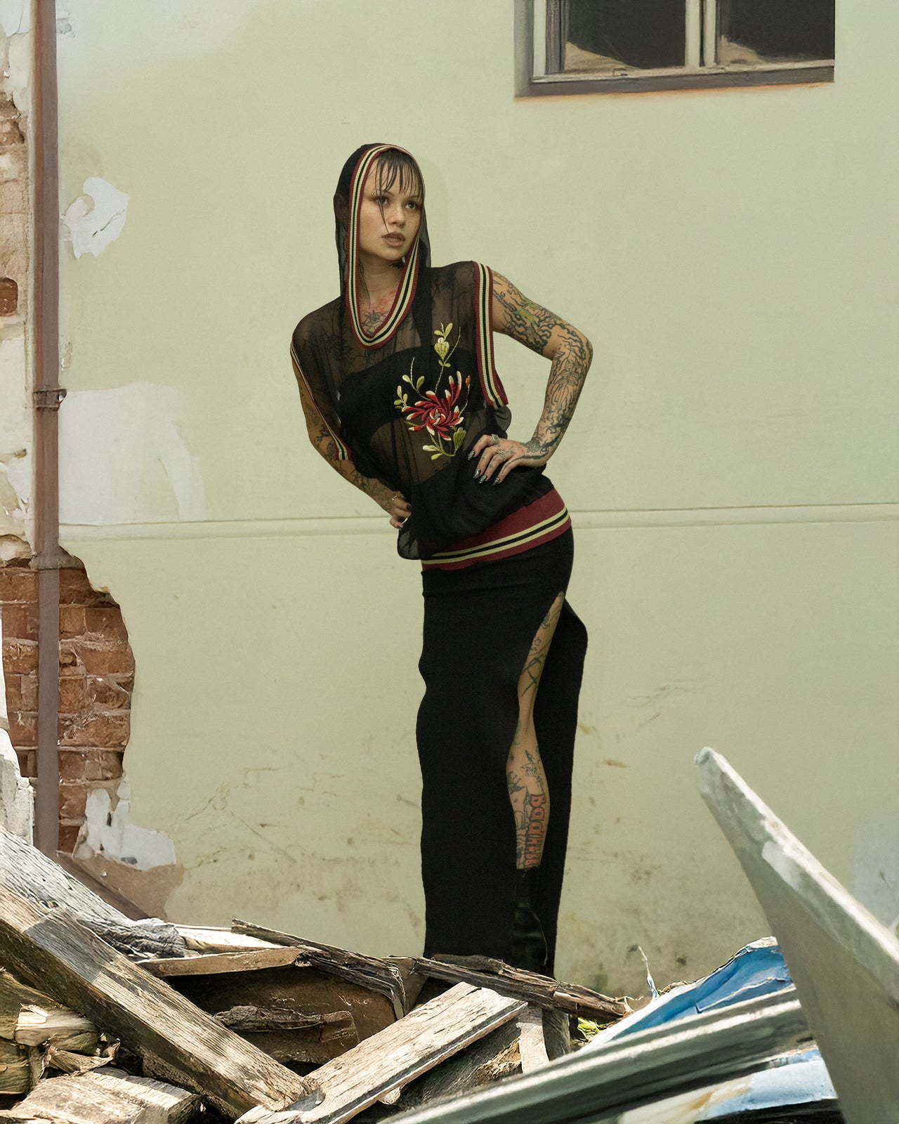 A model wearing a hooded mesh Jean Paul Gaultier basketball tank top with floral motifs and numbers, in a run-down building among debris.