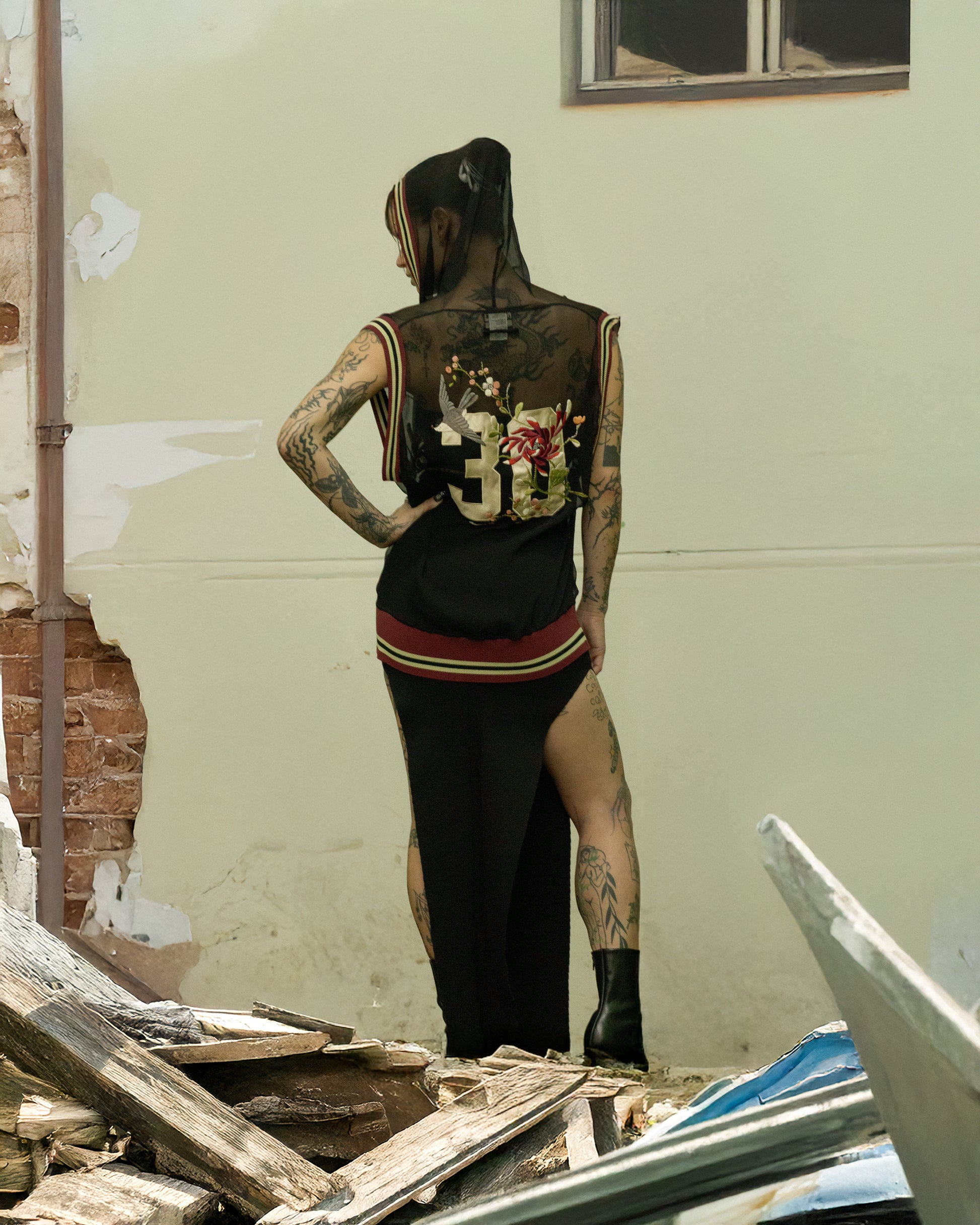 Person wearing a black Jean Paul Gaultier sleeveless top with floral design and number 35, standing in a dilapidated room.