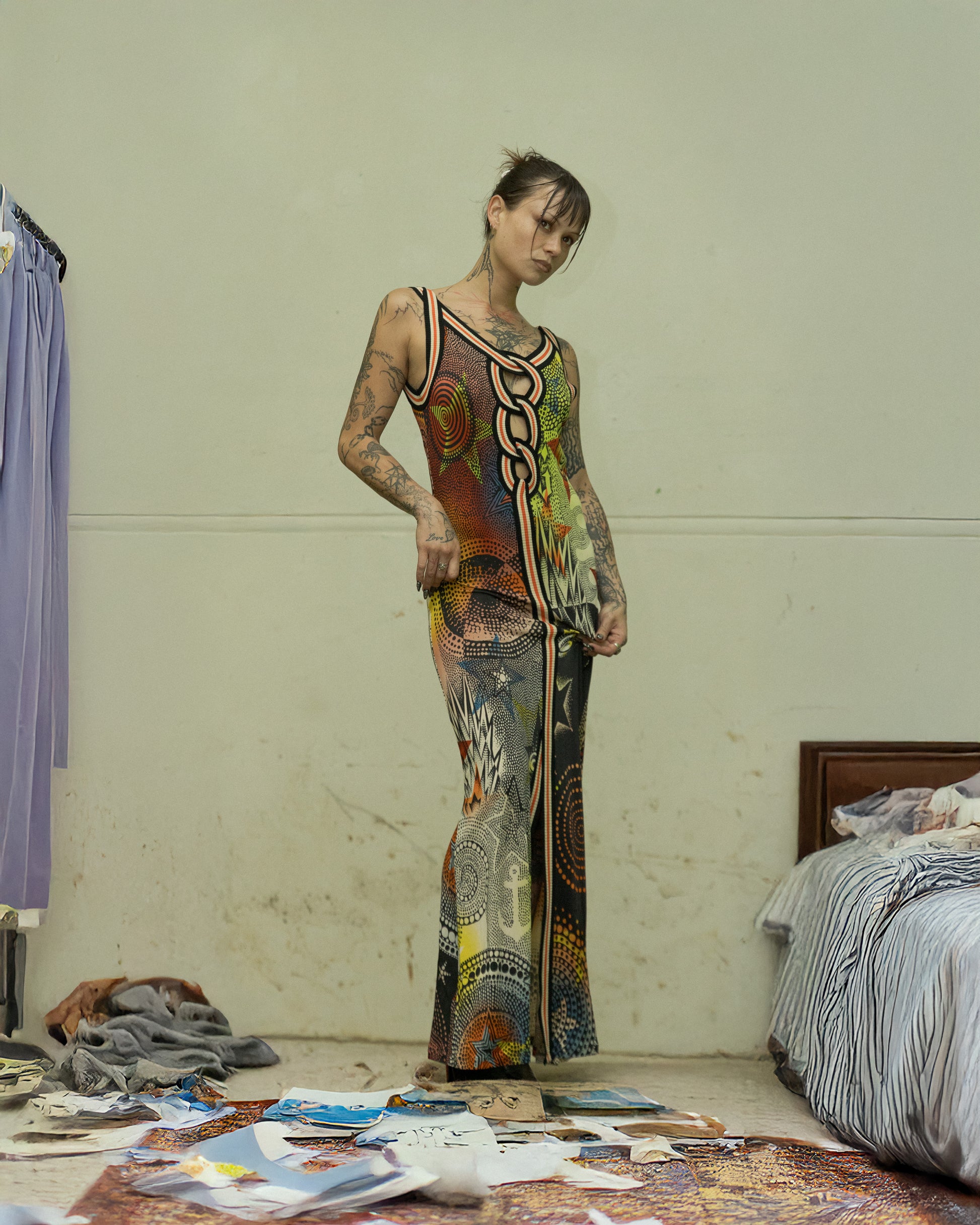 A person standing in a room with a variety of clothes and materials scattered around, wearing a multicoloured Jean Paul Gaultier sleeveless dress with a geometric star print.