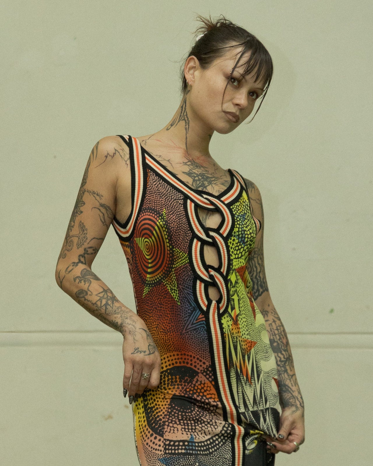 A person standing in a room with a variety of clothes and materials scattered around, wearing a multicoloured Jean Paul Gaultier sleeveless dress with a geometric star print.