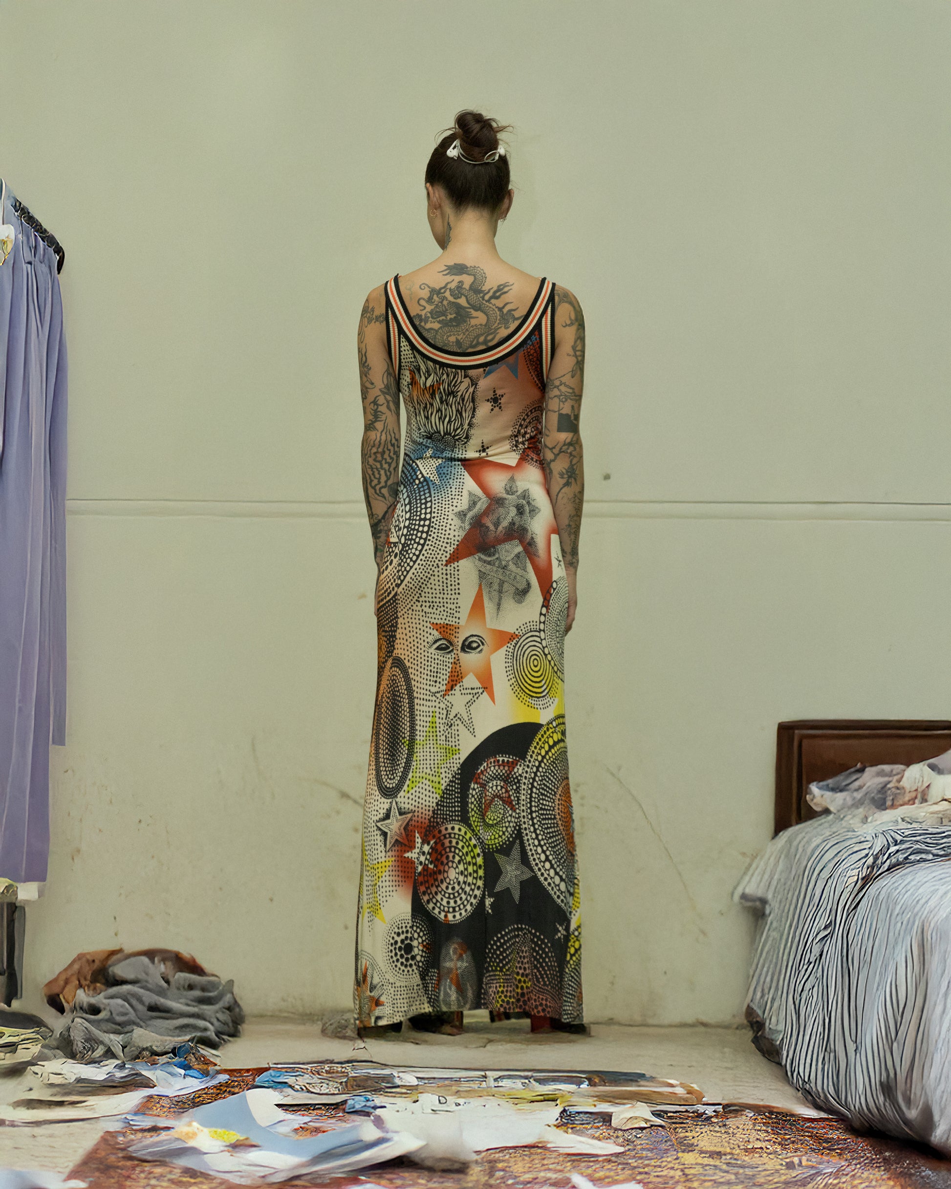 Person wearing a long, patterned Jean Paul Gaultier dress with tattoos visible on arms and back, standing in a room with a bed and clothes.