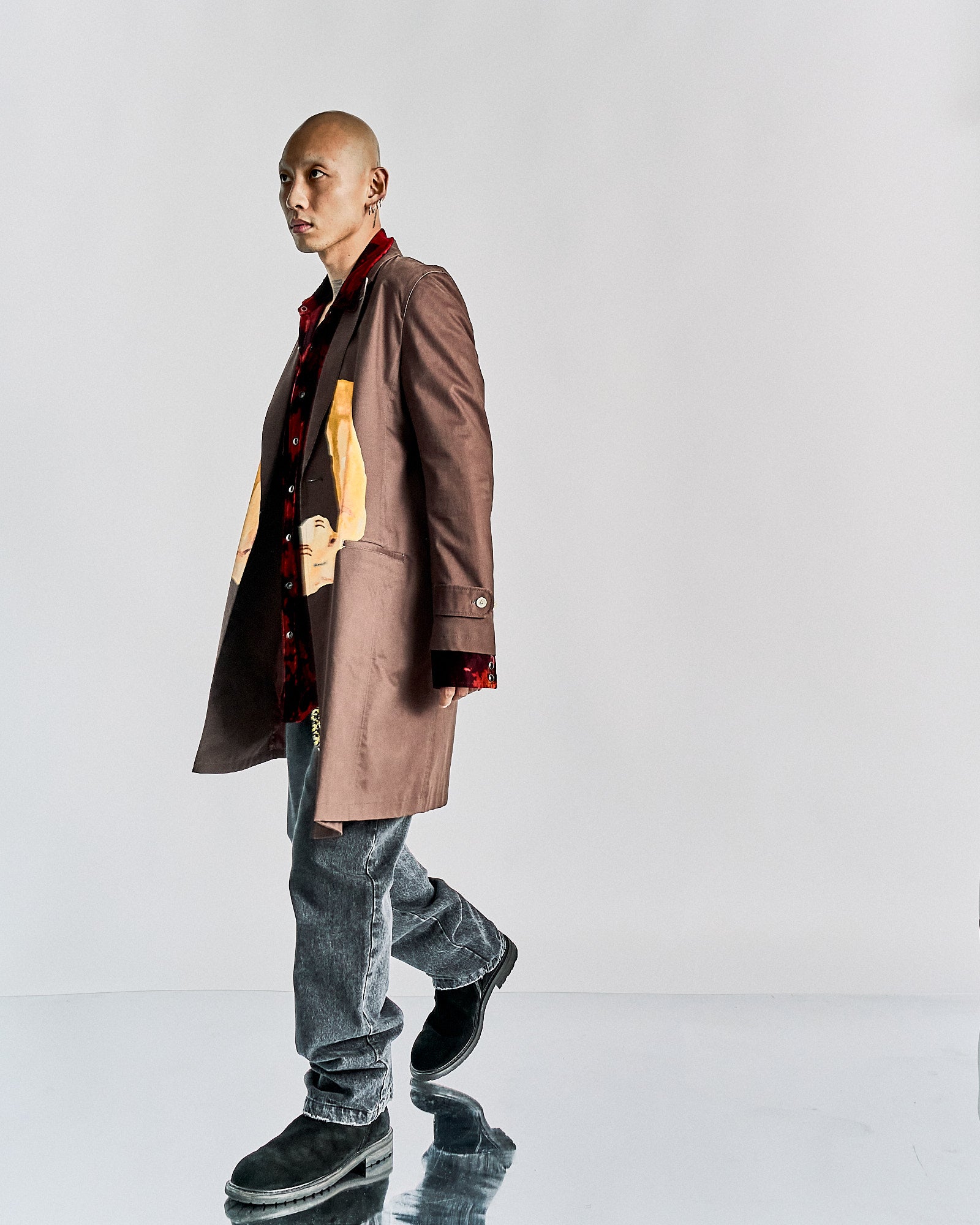 Man in brown John Undercover coat with colourful print, red patterned shirt, and grey jeans. He stands on a reflective surface against a plain white background. The tone is modern and stylish.
