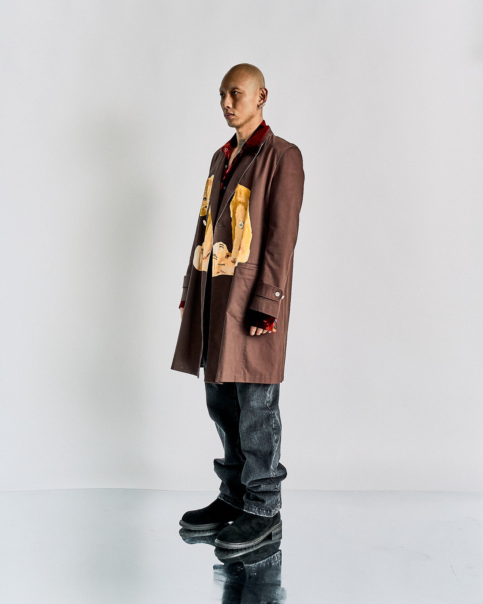A person stands confidently wearing a brown John Undercover coat with artistic patches, over a red shirt. They pair it with grey jeans and black boots