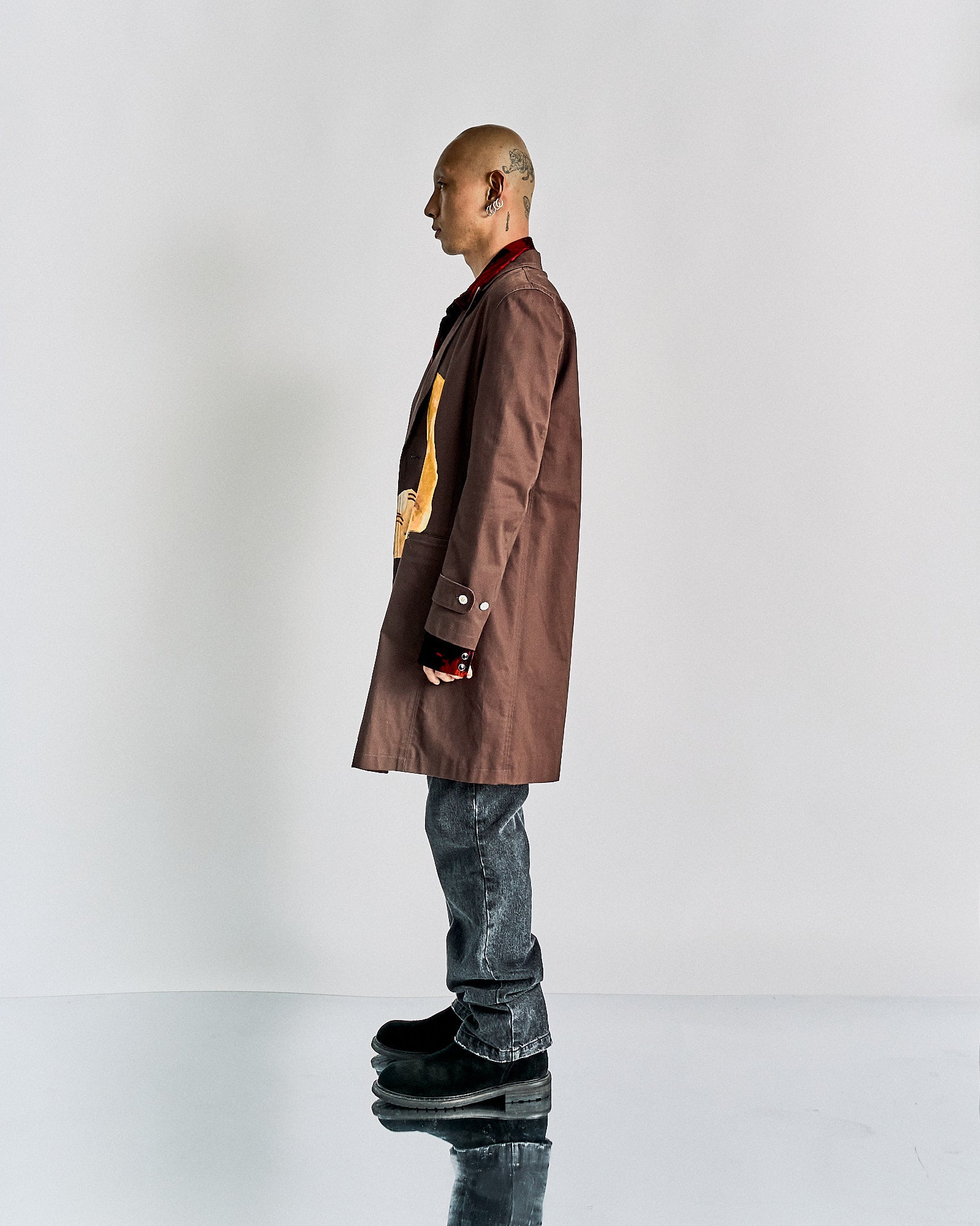 A person stands confidently wearing a brown John Undercover coat with artistic patches, over a red shirt. They pair it with grey jeans and black boots